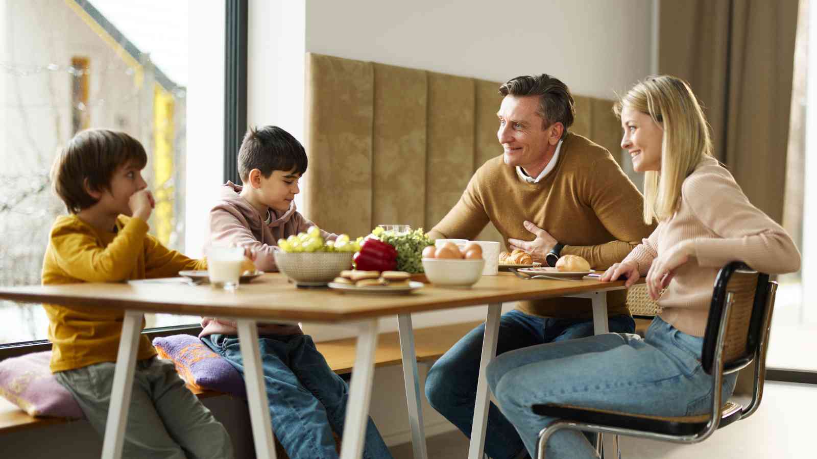 A family of four sits at a dining table with food and drinks, smiling and talking together in a bright room.