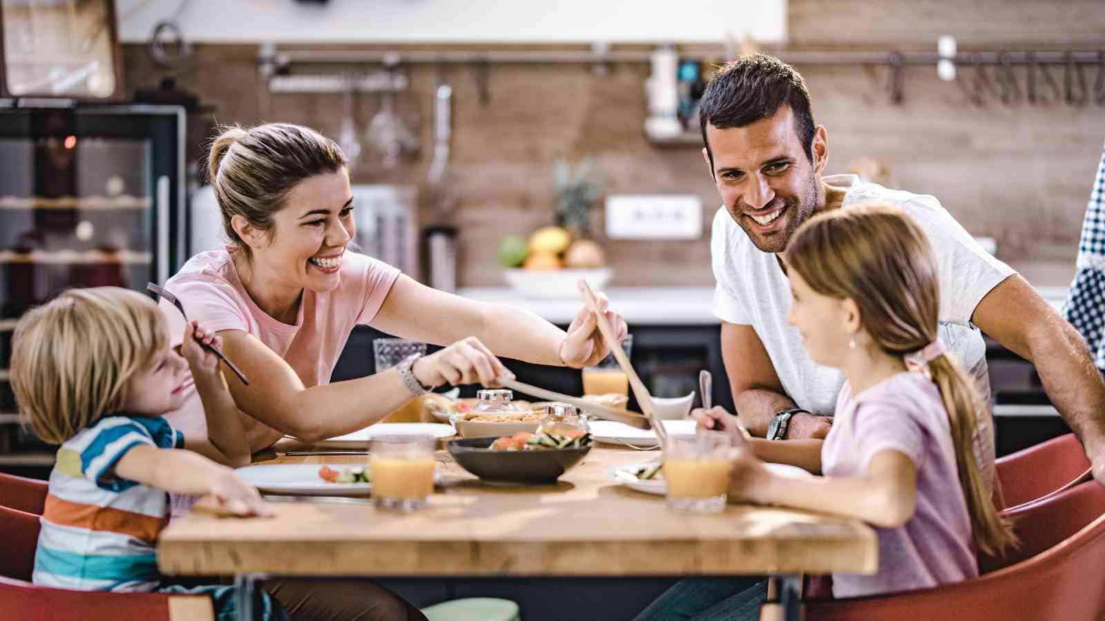 A family of four sits at a kitchen table, smiling and eating together, with plates of food and glasses of juice in front of them.
