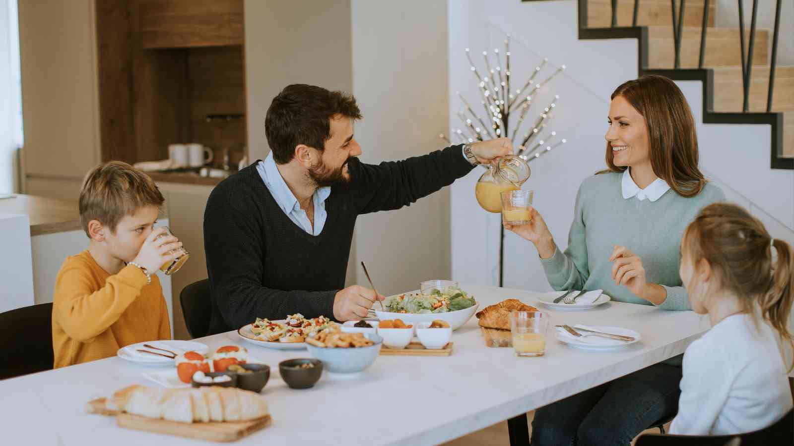 A family of four sits at a dining table eating breakfast; an adult pours juice for another while the children drink and eat.