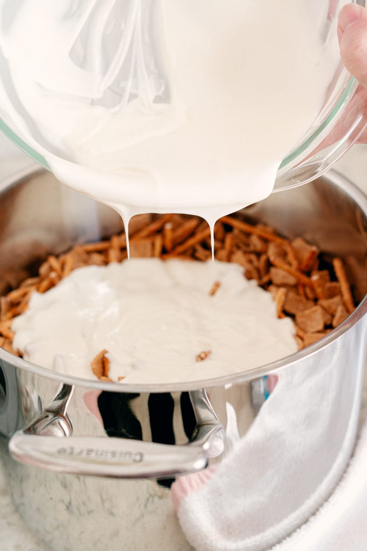 A bowl of white liquid is being poured over a pot filled with pieces of cereal or crackers.