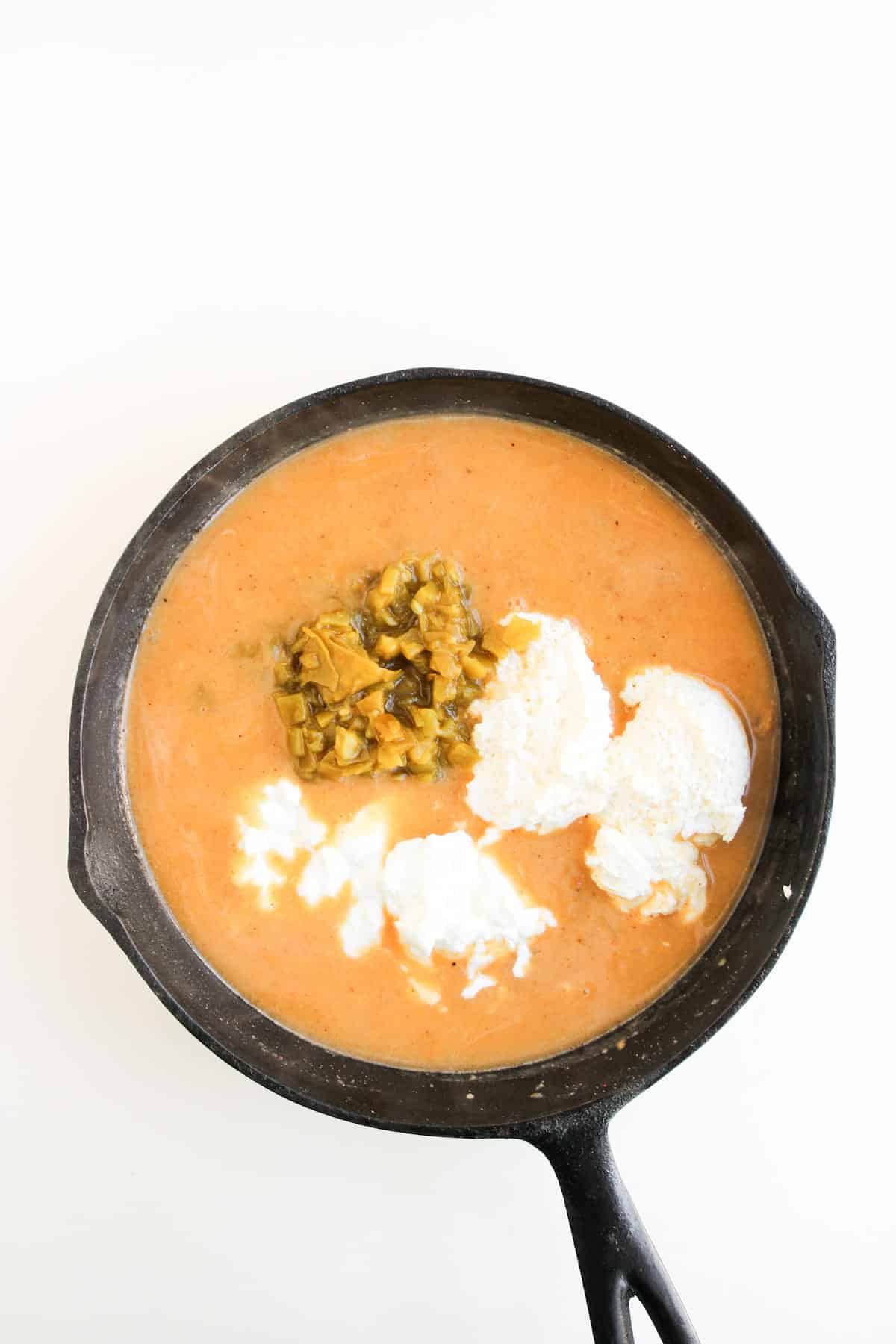 A black skillet filled with soup, topped with white dollops of cream and a portion of chopped chilly vegetables, on a white background.