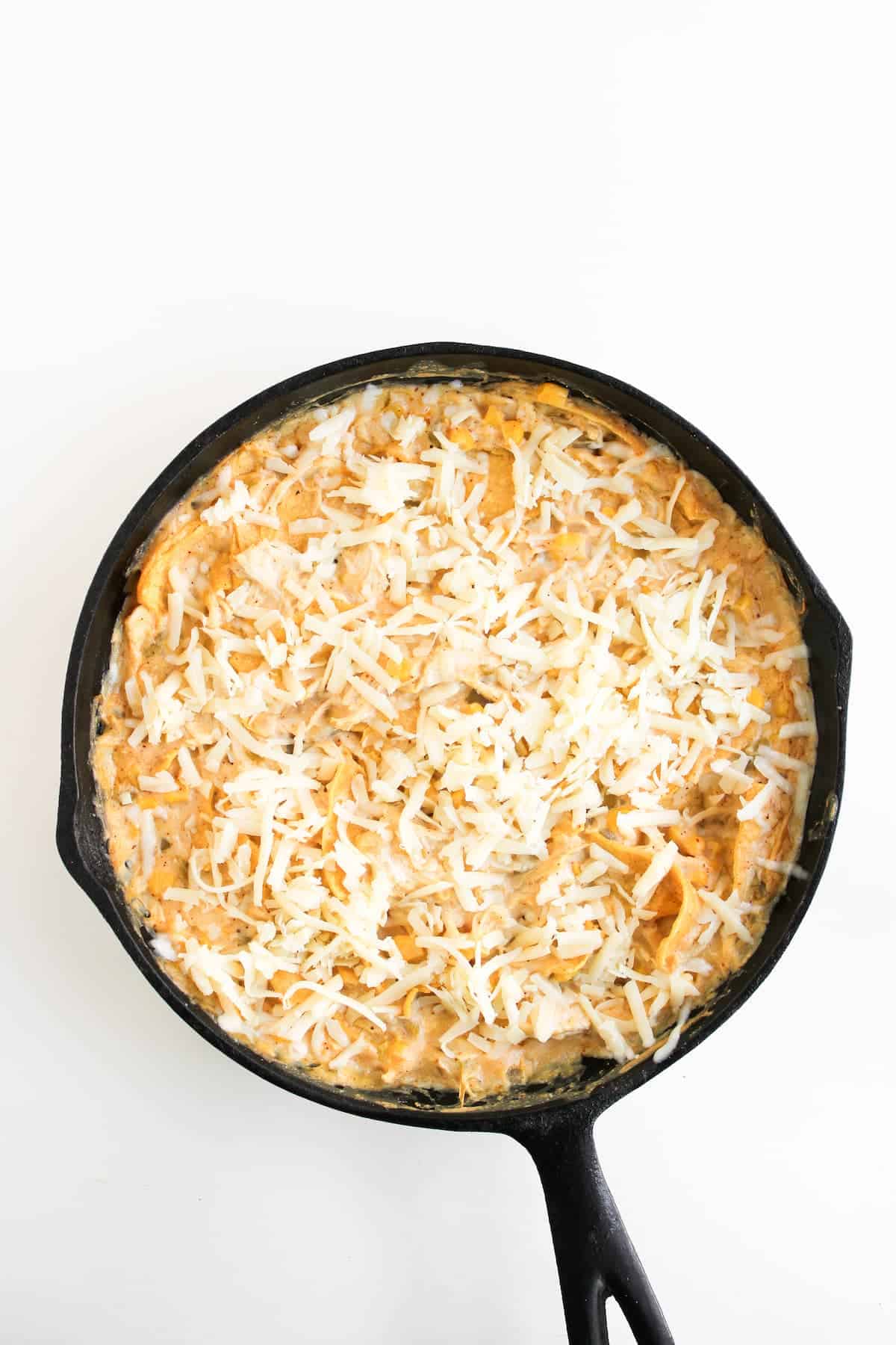 A black cast iron skillet filled with a creamy baked dish topped with shredded cheese, set against a white background.