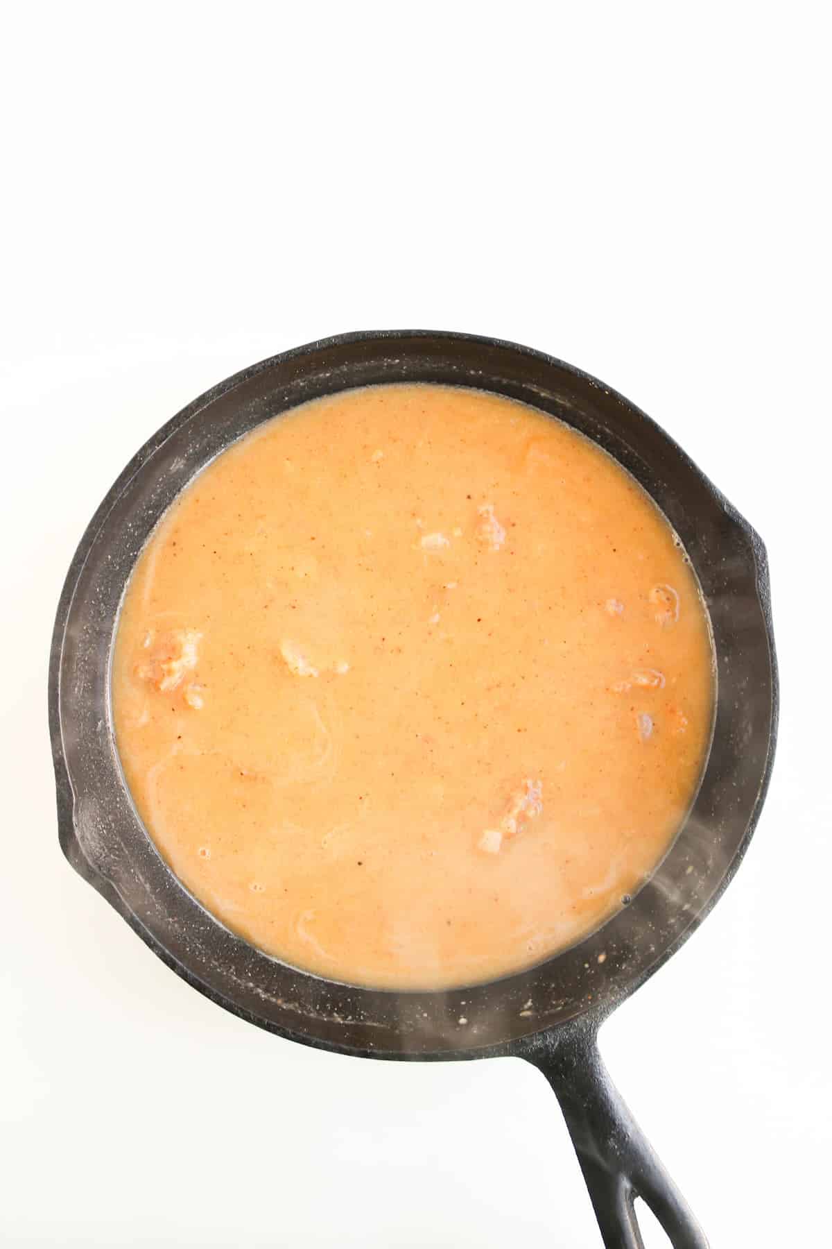 A black cast iron skillet containing a light brown, creamy sauce with small chunks, set against a white background.