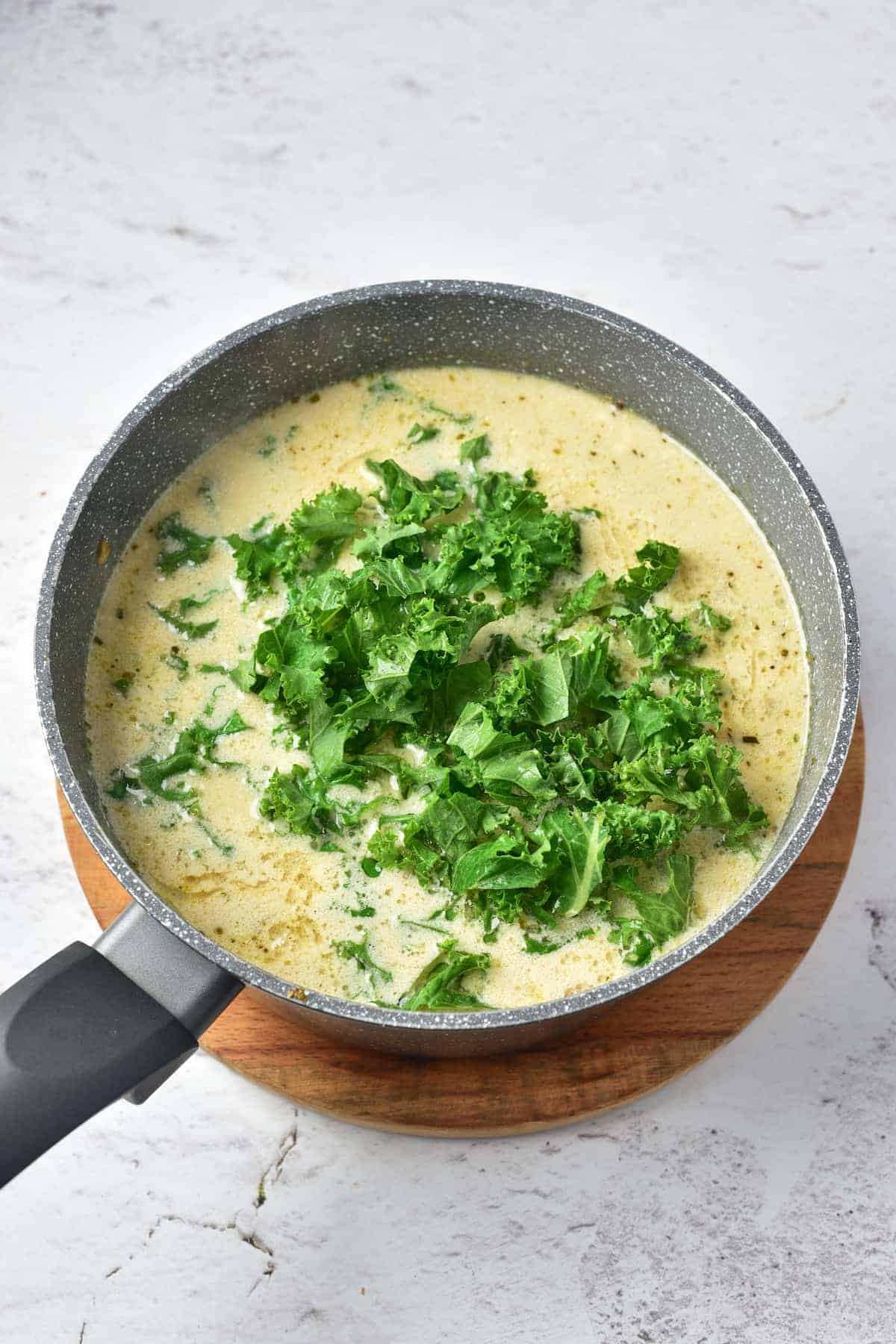 A pot of creamy soup with chopped kale on top, sitting on a round wooden trivet against a light, textured background.