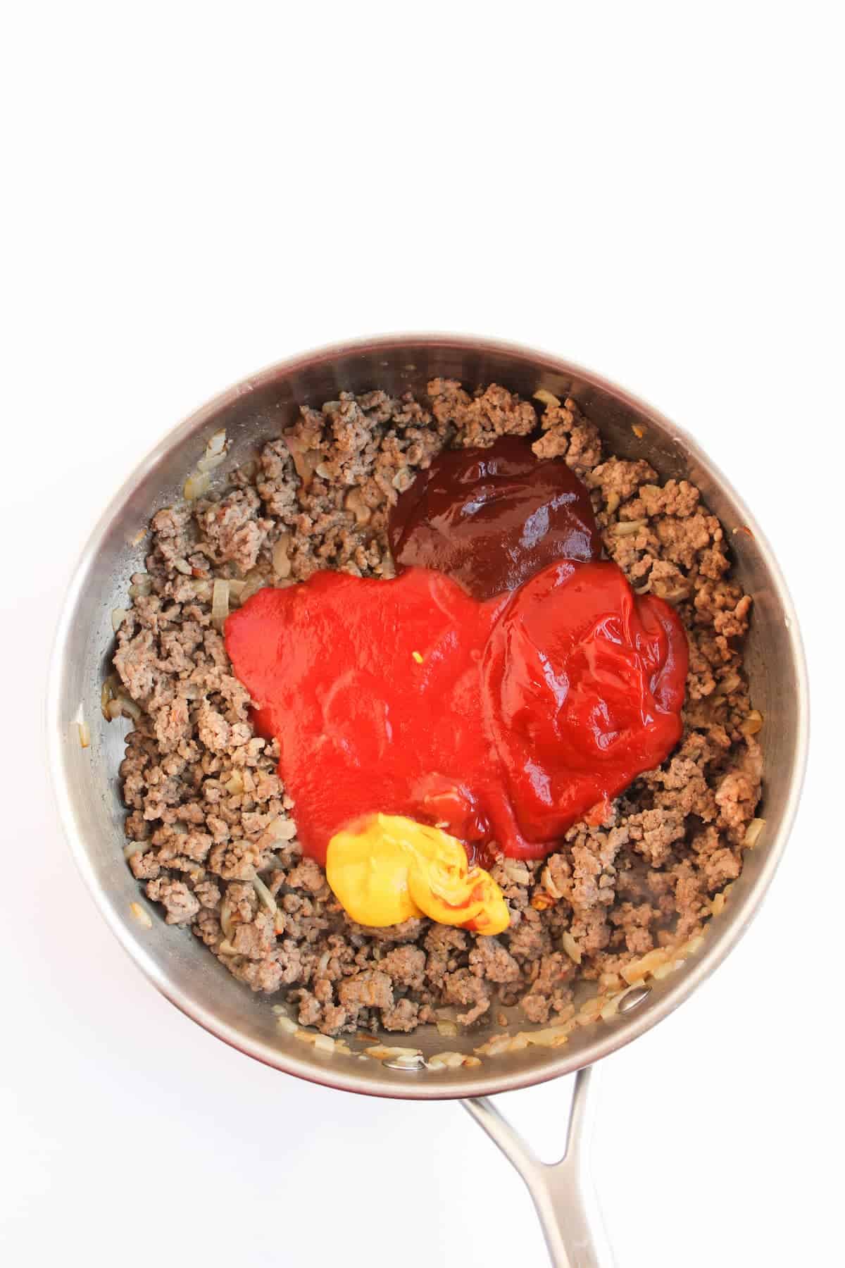 Ground beef and onions cooking in a pan with ketchup, tomato sauce, barbecue sauce, and mustard added on top.