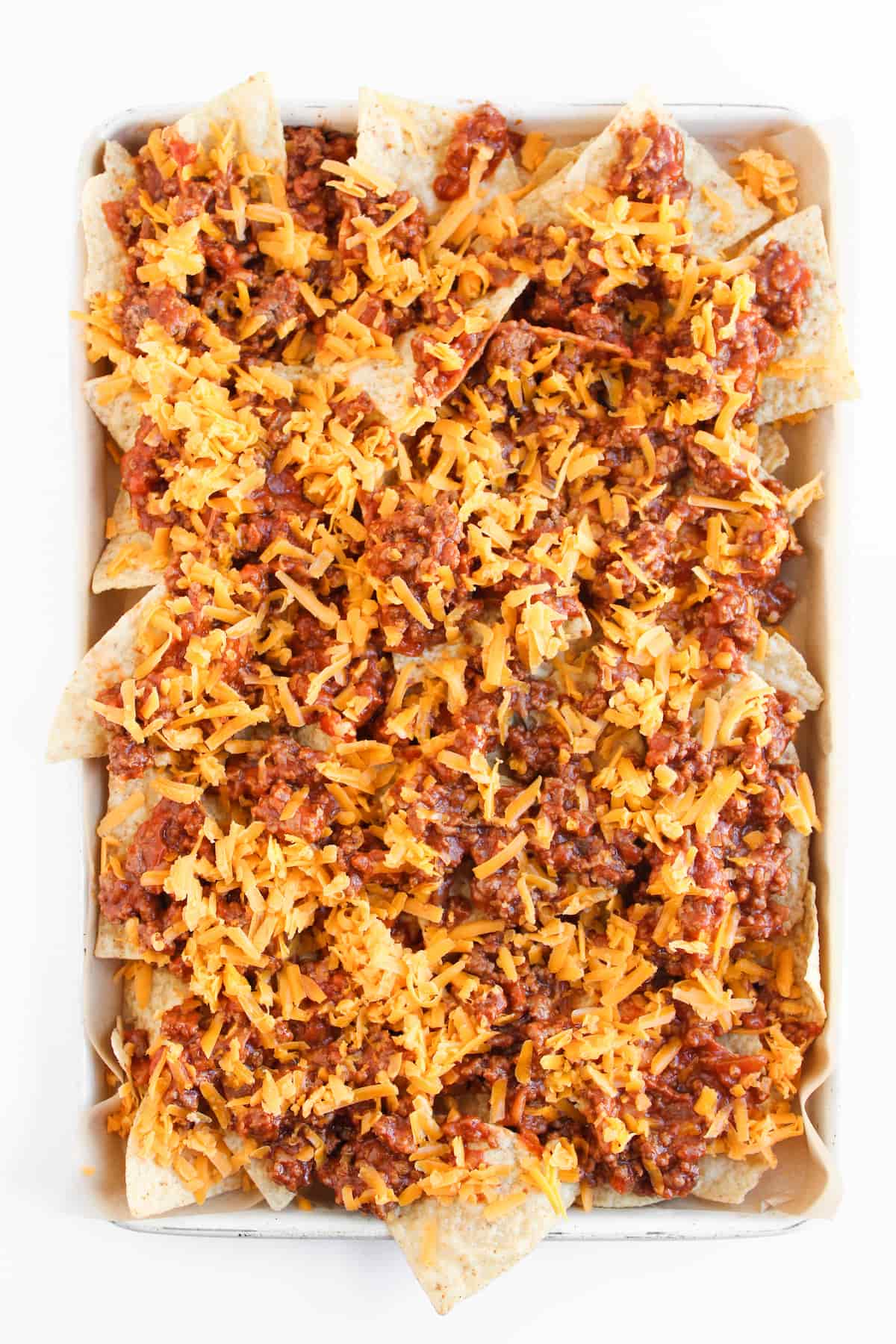 A baking sheet filled with tortilla chips topped with cooked ground beef and shredded cheddar cheese.