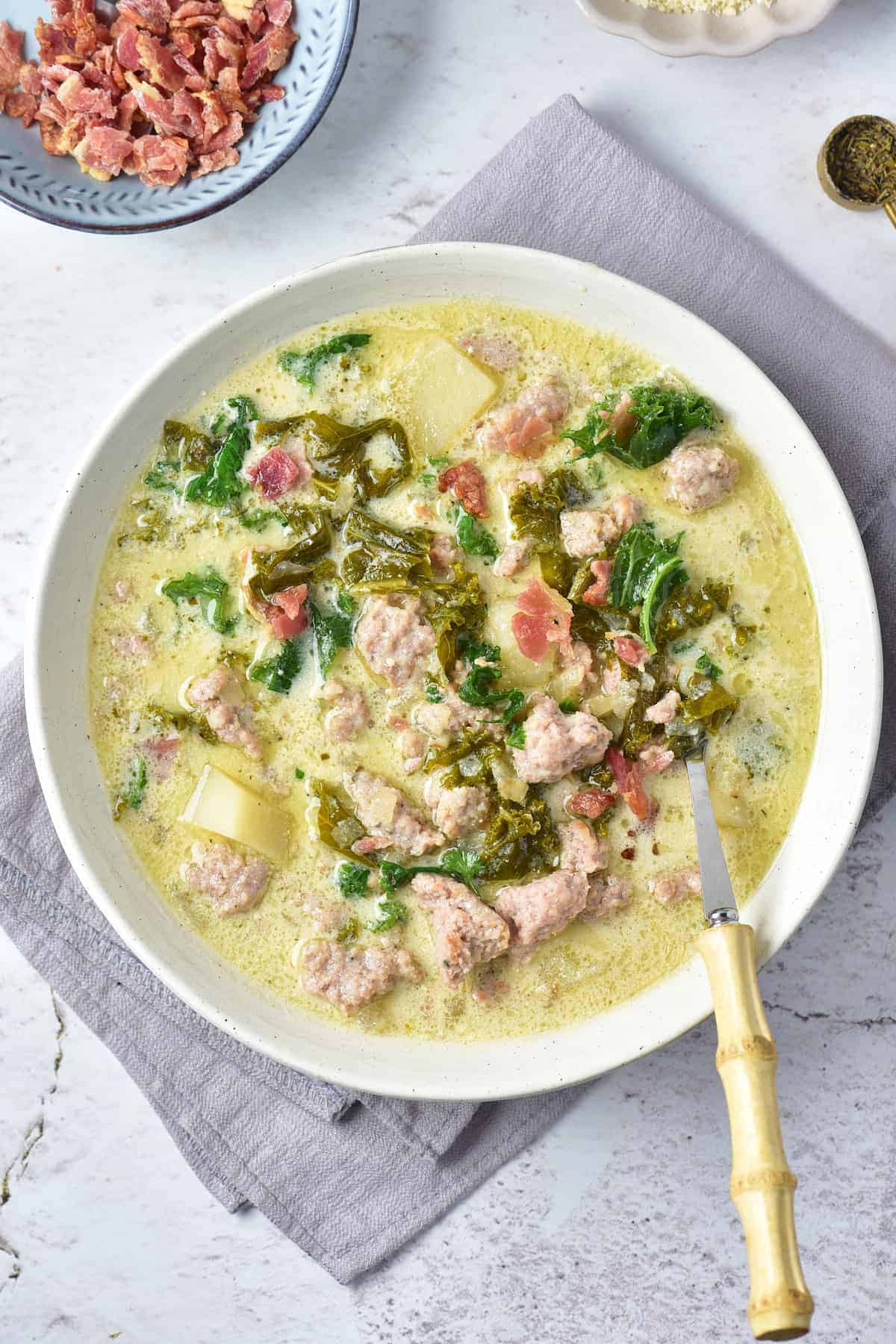 A bowl of creamy soup with sausage, kale, potatoes, and bacon bits, placed on a gray napkin with a spoon and a small bowl of bacon pieces nearby.