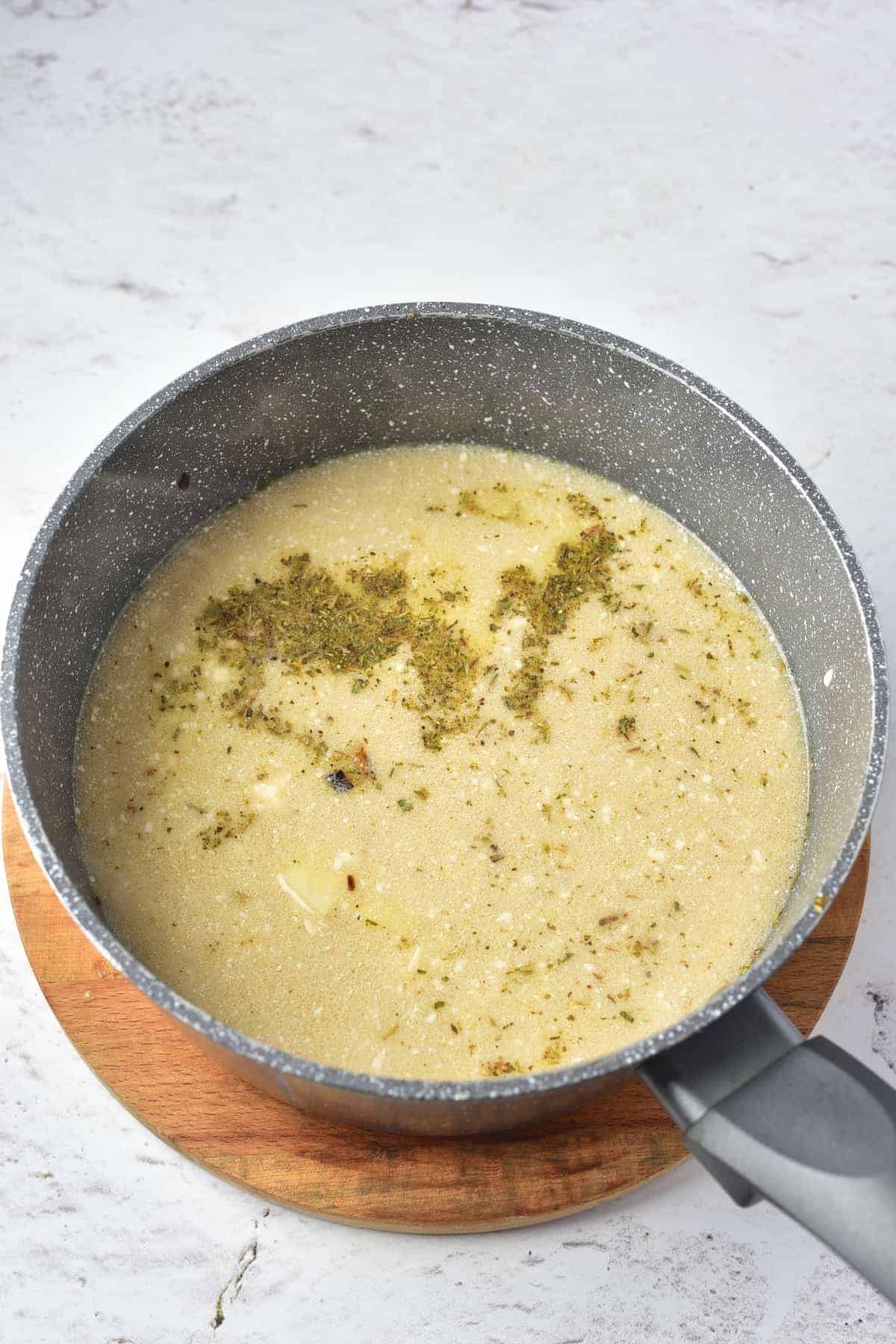 A saucepan filled with a light-colored soup or broth, topped with dried herbs, sits on a round wooden trivet against a light background.