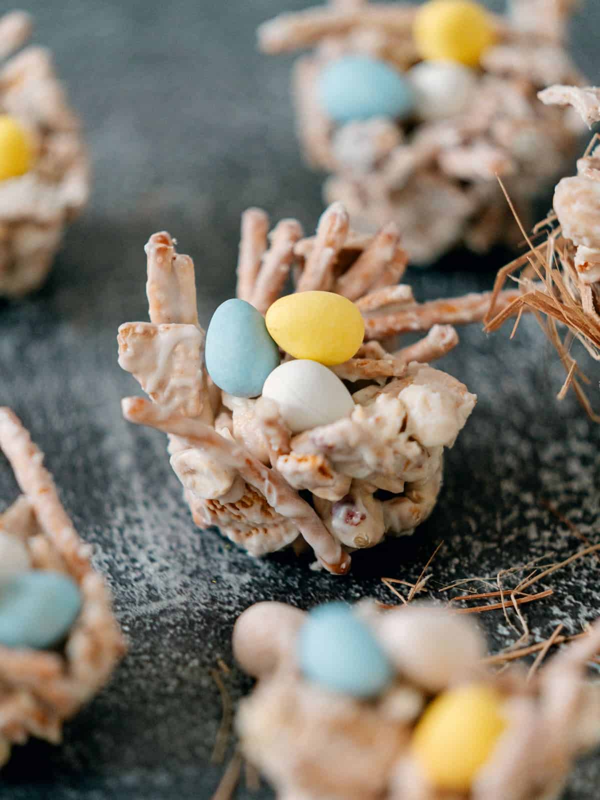 A close-up of a treat made from crunchy sticks and marshmallows shaped like a nest, topped with pastel candy eggs. Other similar treats are visible in the background.