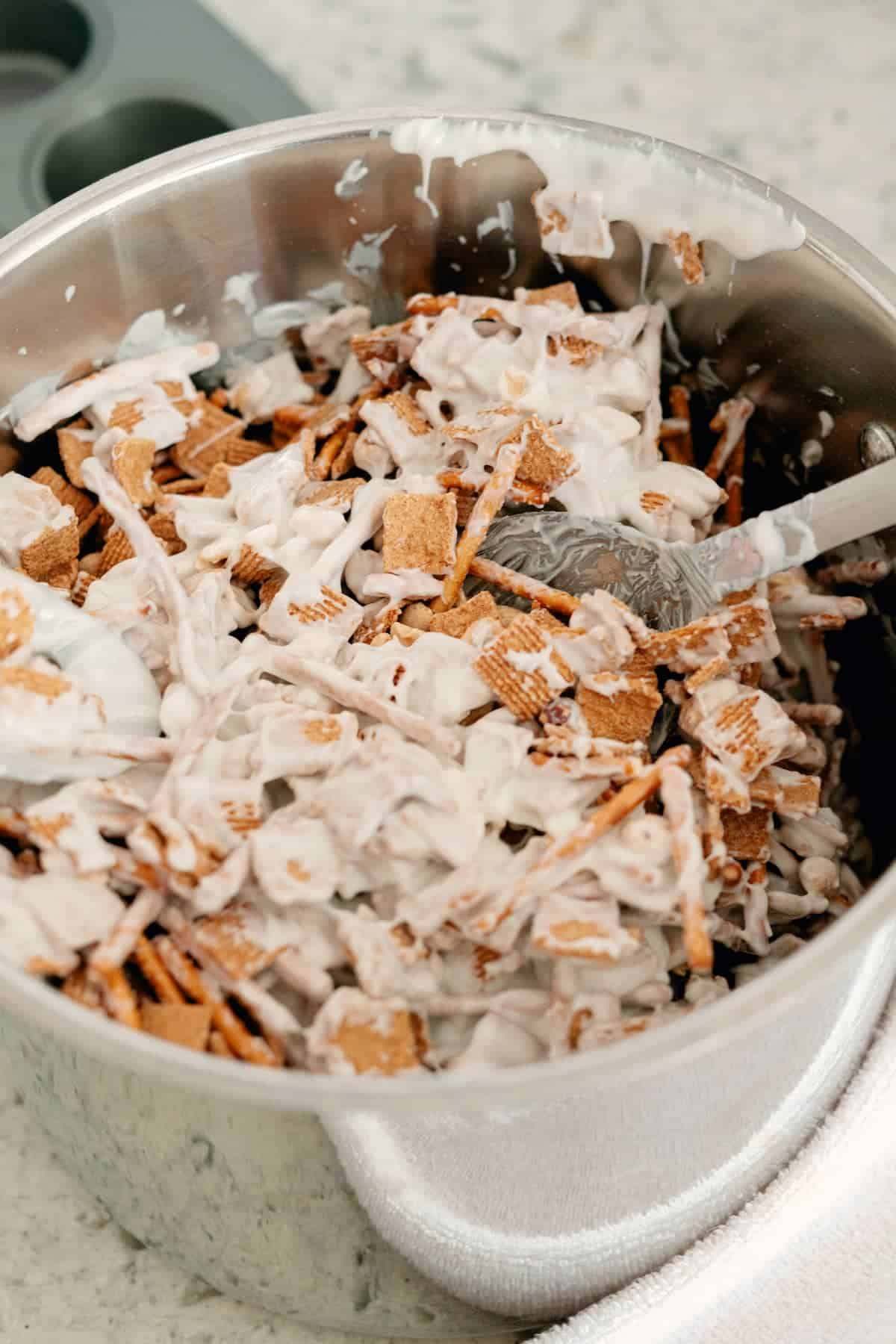 A metal bowl filled with a mixture of cereal pieces and melted white coating, being stirred with a metal spoon.