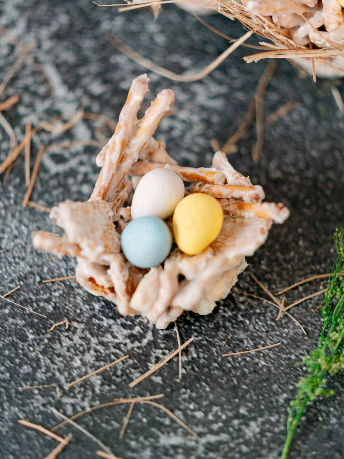 A small edible nest made of pretzel sticks and a white coating holds three candy eggs in white, yellow, and blue, placed on a gray textured surface.
