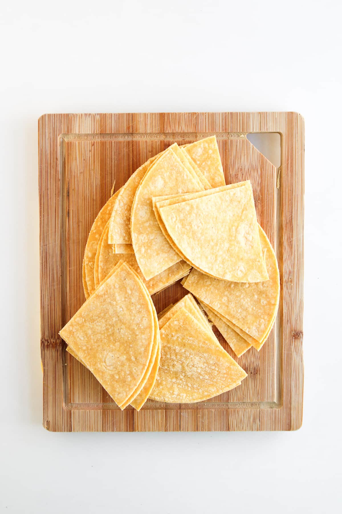 Several corn tortillas are stacked and cut into quarters, arranged on a wooden cutting board against a white background.