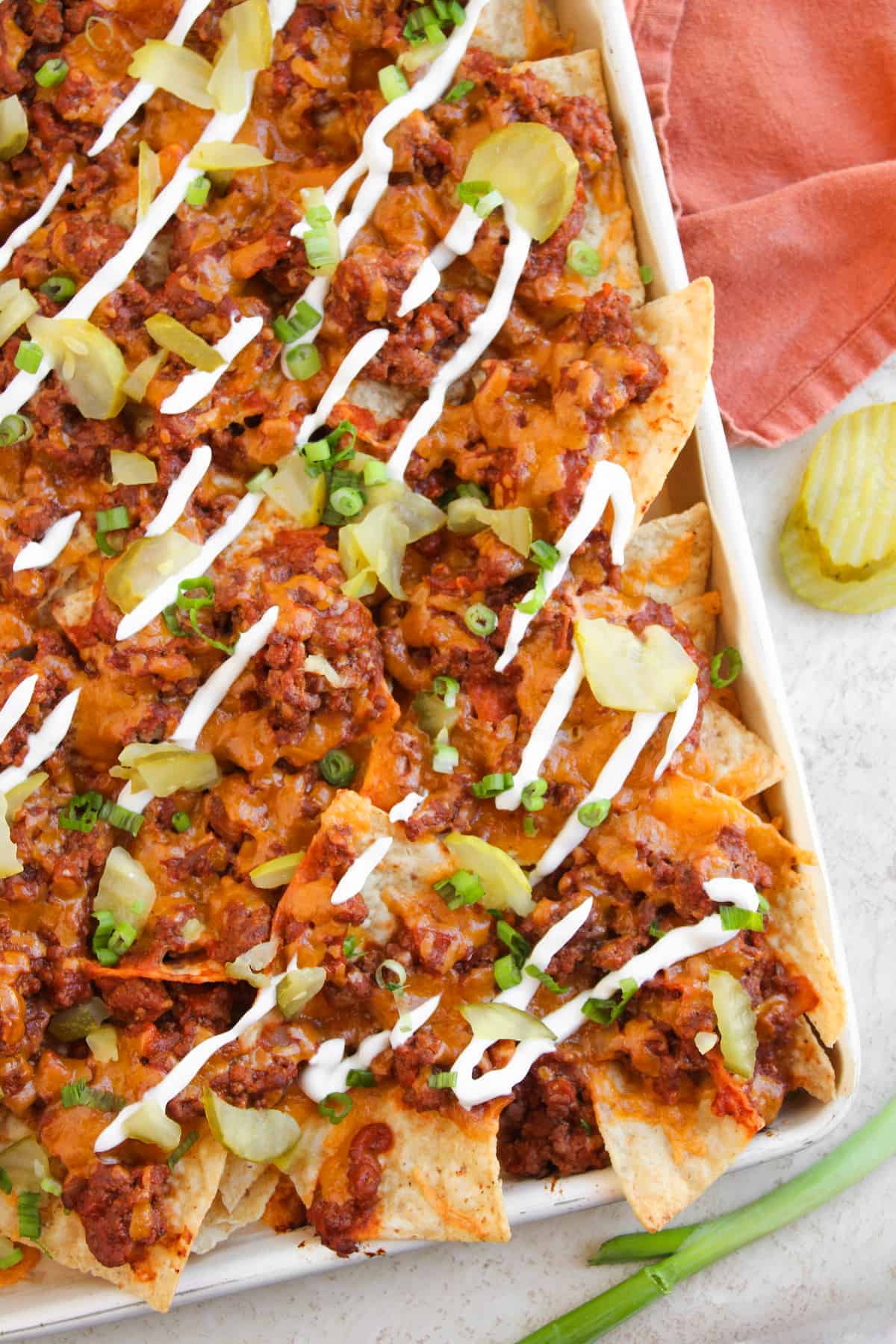 A tray of nachos topped with ground meat, melted cheese, sliced pickles, chopped green onions, and drizzled with sour cream. An orange cloth and pickle slices are nearby.