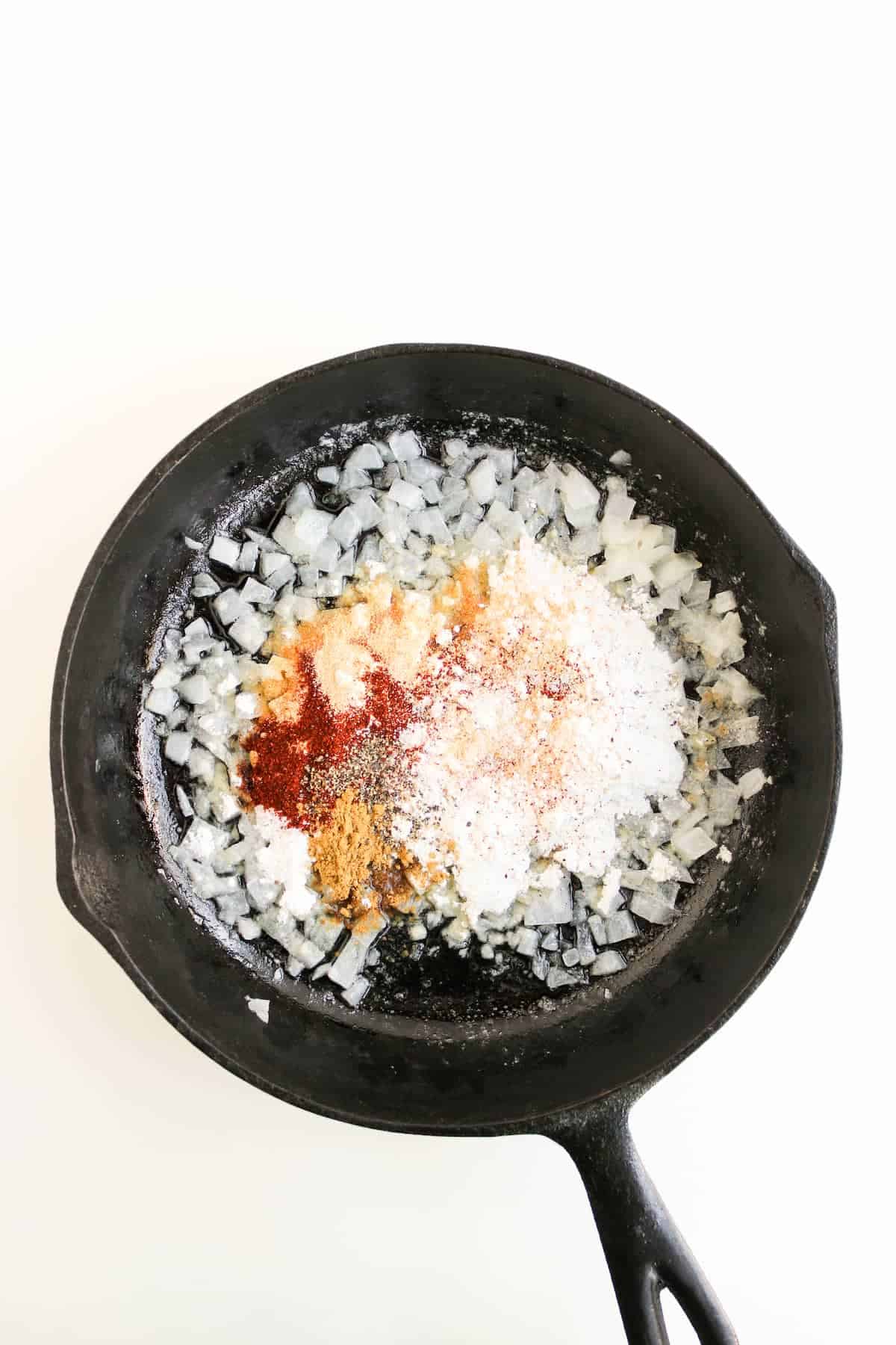 Chopped onions and various powdered spices in a black cast iron skillet on a white background.
