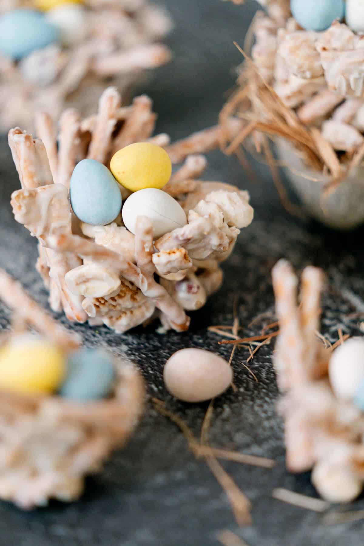 Edible nests made of coated pretzel sticks, filled with pastel-colored candy eggs, arranged on a dark surface with straw-like decorations.