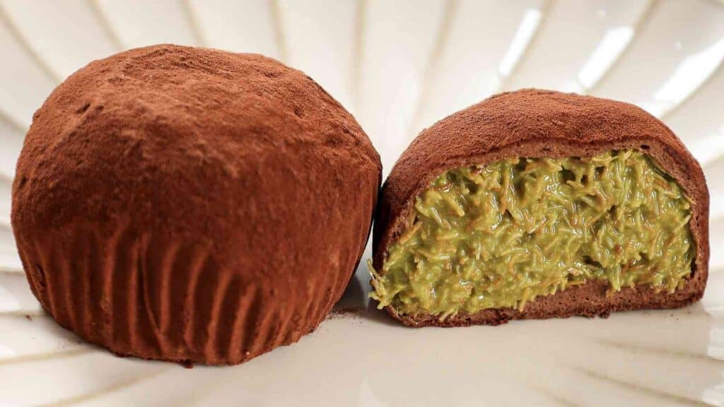 Two chocolate-coated truffles on a plate, one whole and one cut in half to show a green, textured filling inside.