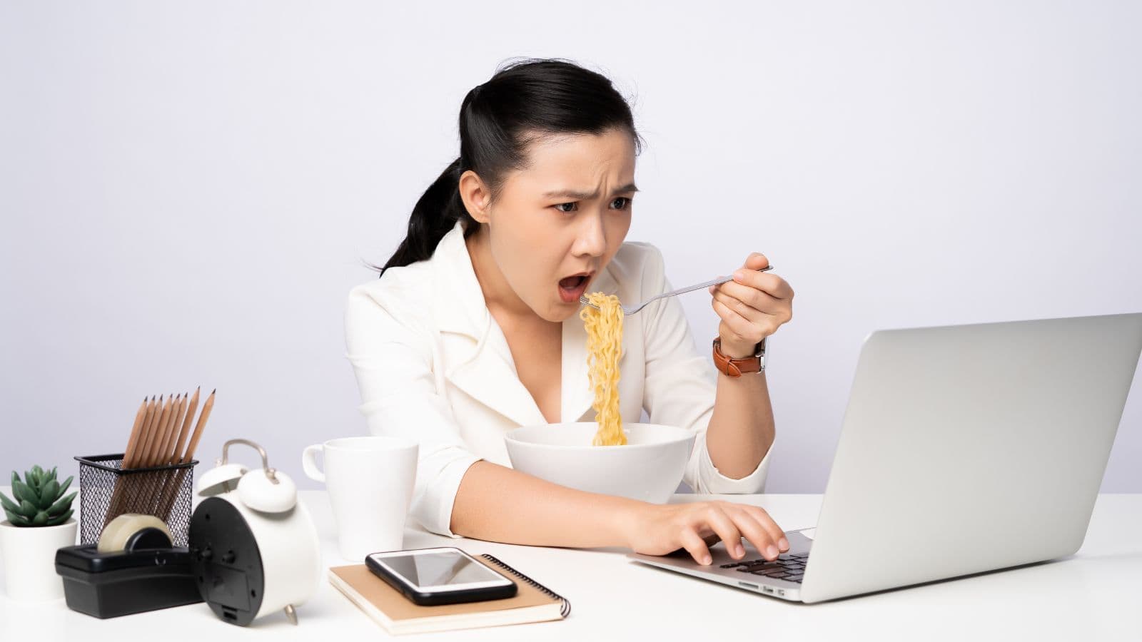 A woman eating noodles in front of a laptop appears surprised while working at a desk with office supplies.