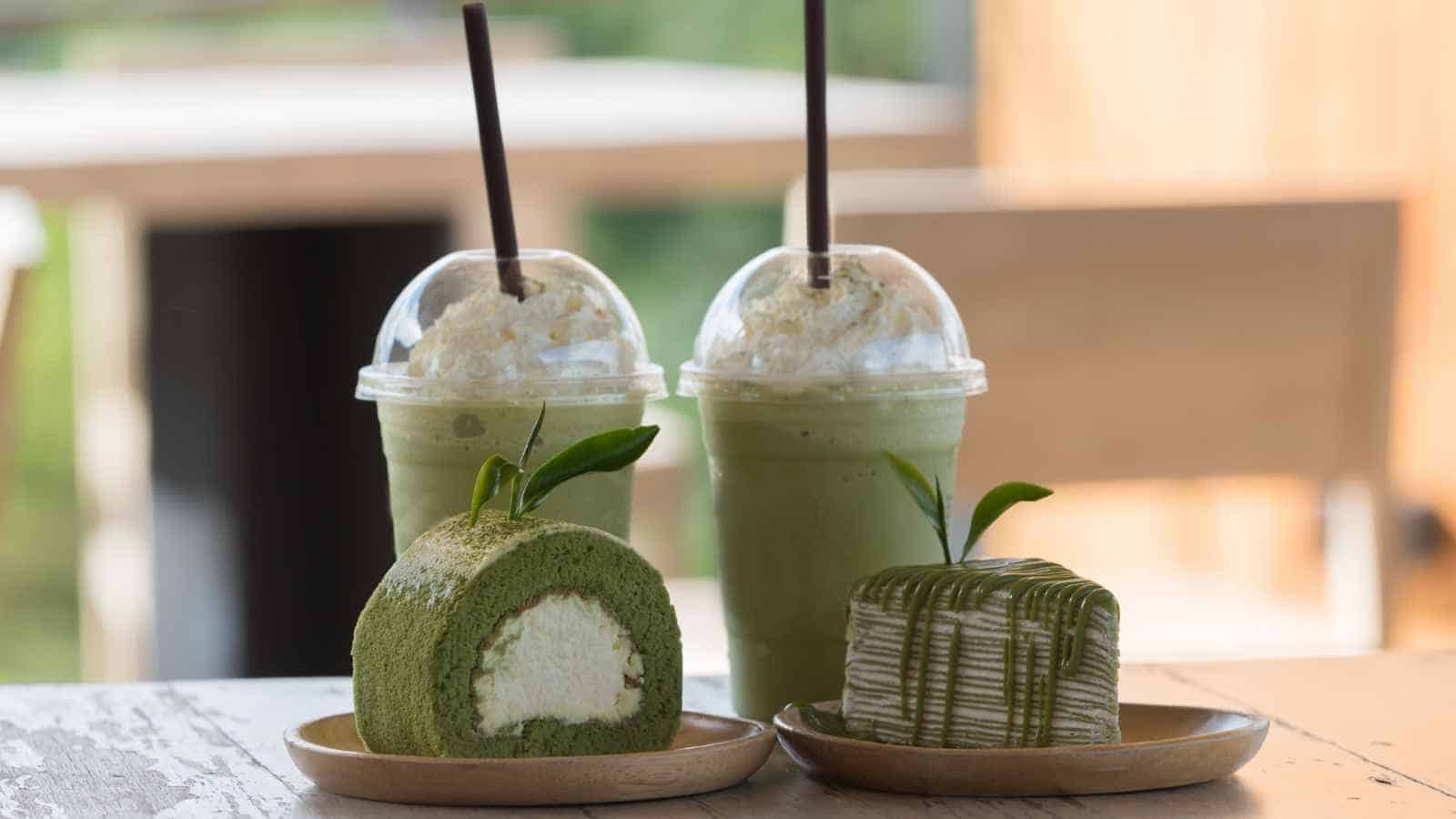 Two green tea-flavored drinks with whipped cream and straws behind a matcha Swiss roll and a matcha cake on wooden plates, all on a light wooden table.