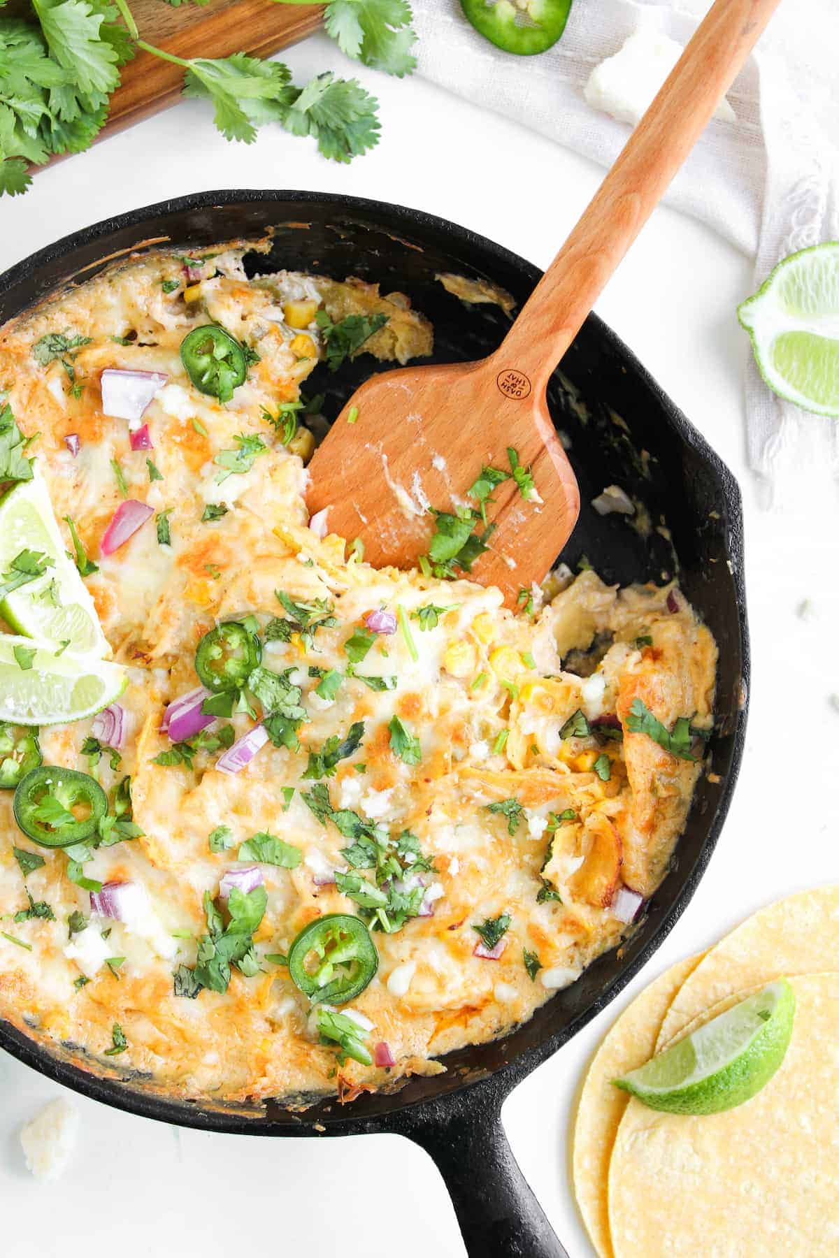 A skillet of cheesy white chicken enchilada topped with sliced jalapeños, chopped red onion, cilantro, and lime wedges, with corn tortillas and lime on the side.
