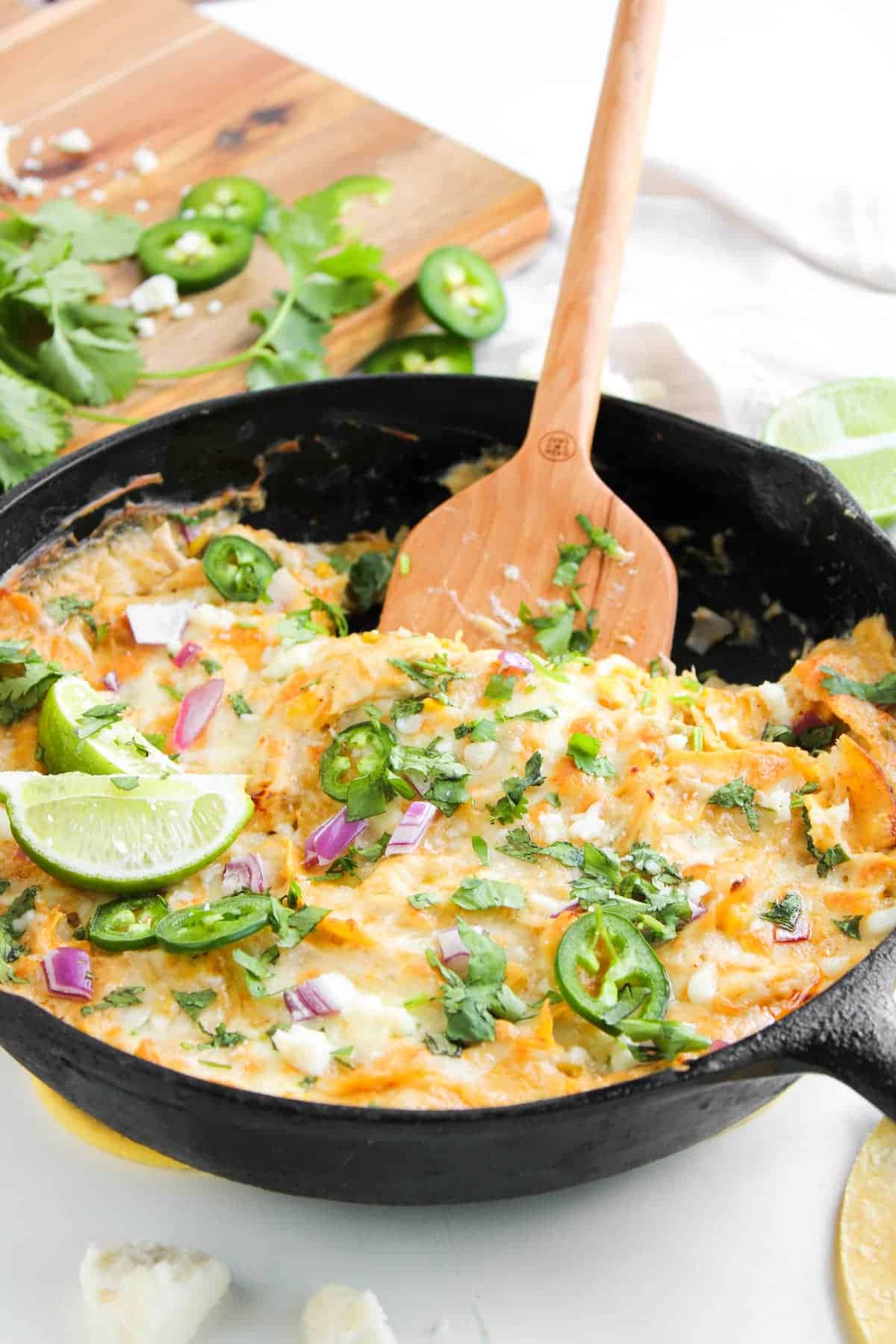 A cast iron skillet filled with cheesy enchilada casserole, topped with jalapeño slices, lime wedges, red onion, and cilantro, with a wooden spoon resting inside.