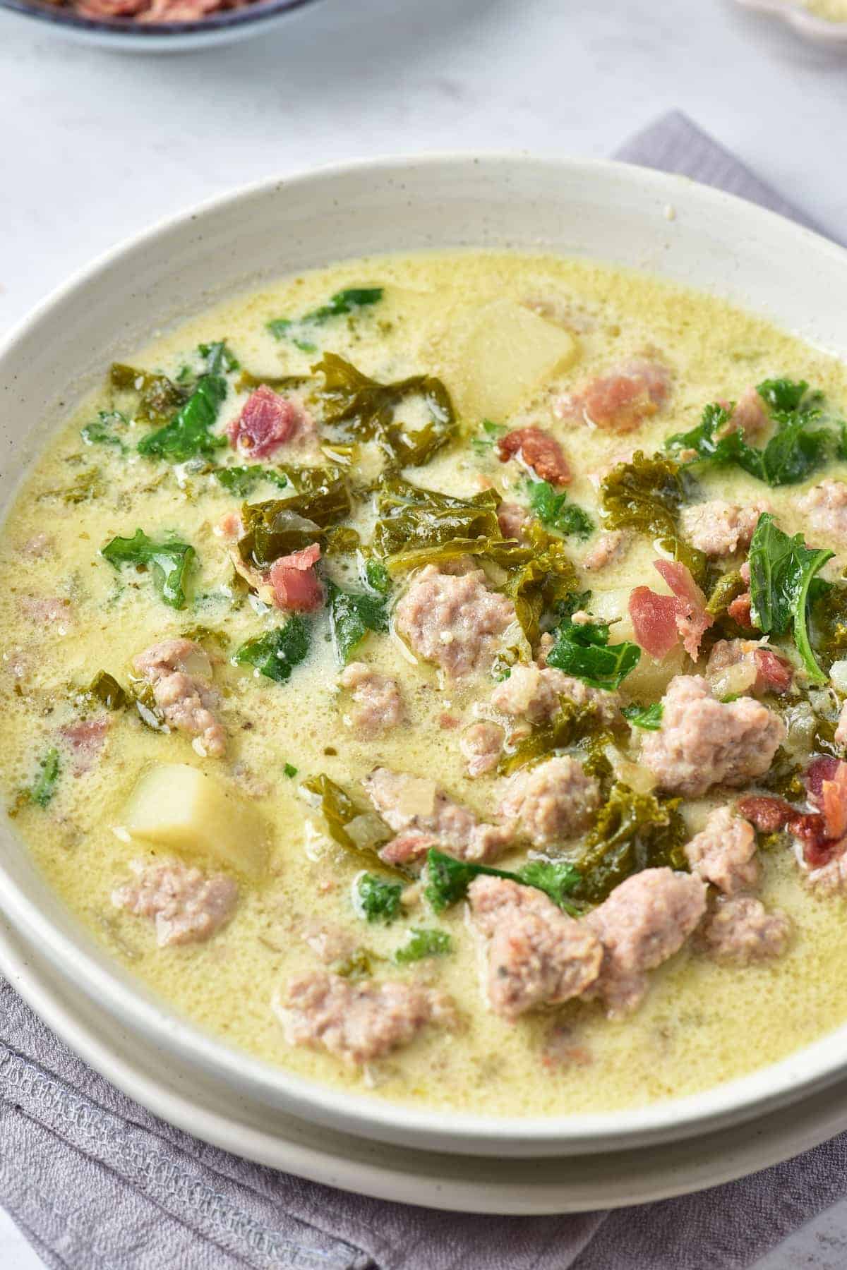 A bowl of creamy soup with sausage, potatoes, kale, and bacon pieces, served on a light-colored napkin.