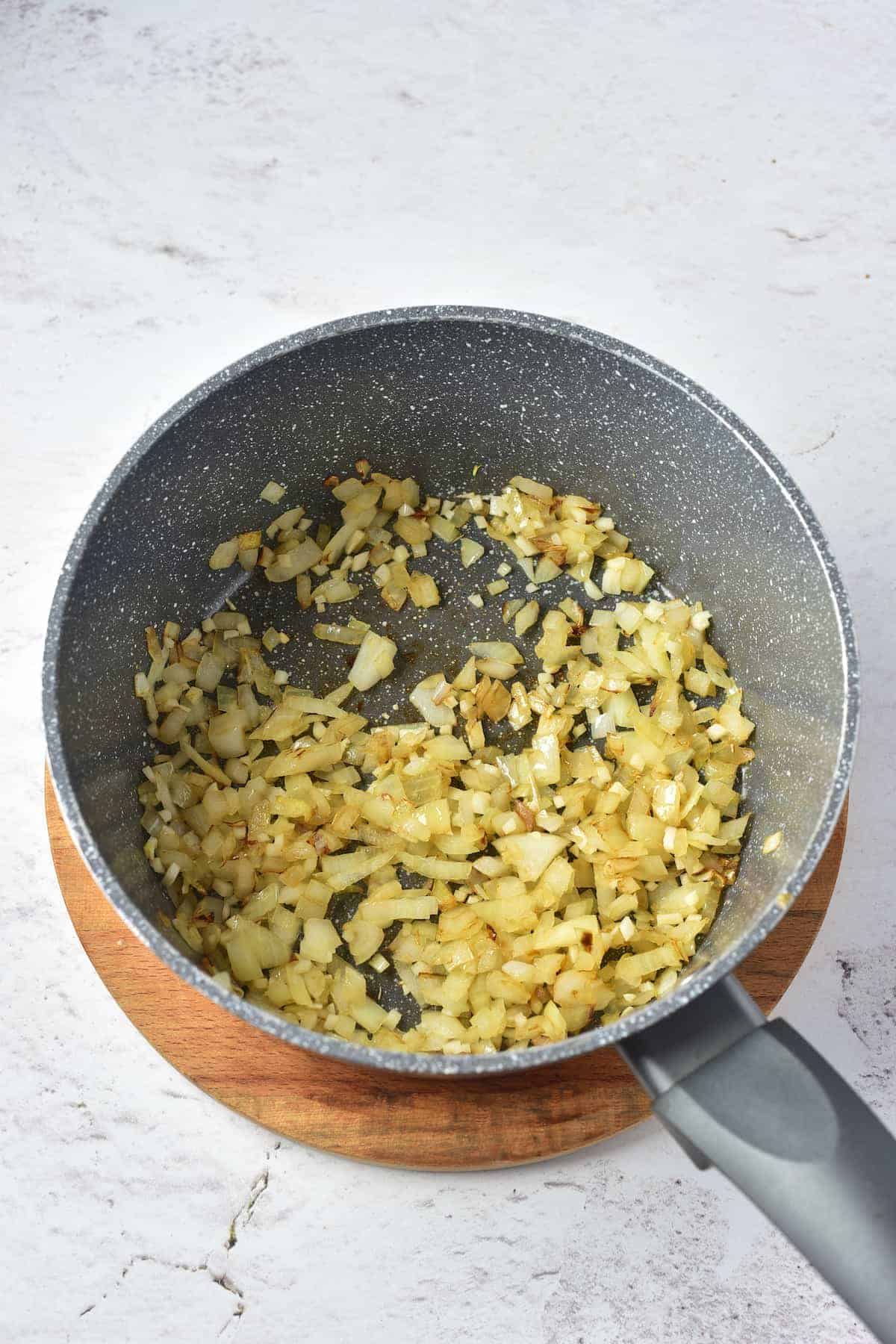Chopped onions being sautéed in a nonstick saucepan placed on a wooden trivet.