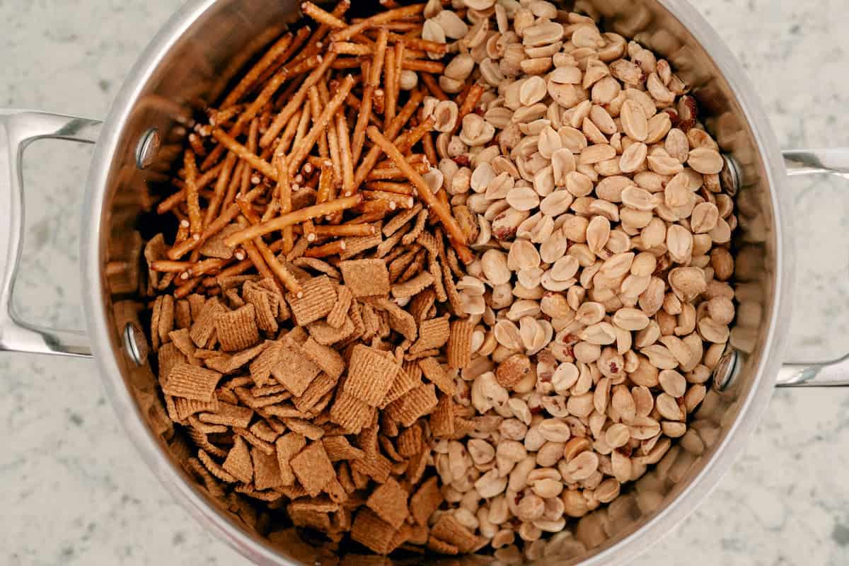 A metal pot containing pretzel sticks, plain peanuts, and square cereal pieces on a light countertop.