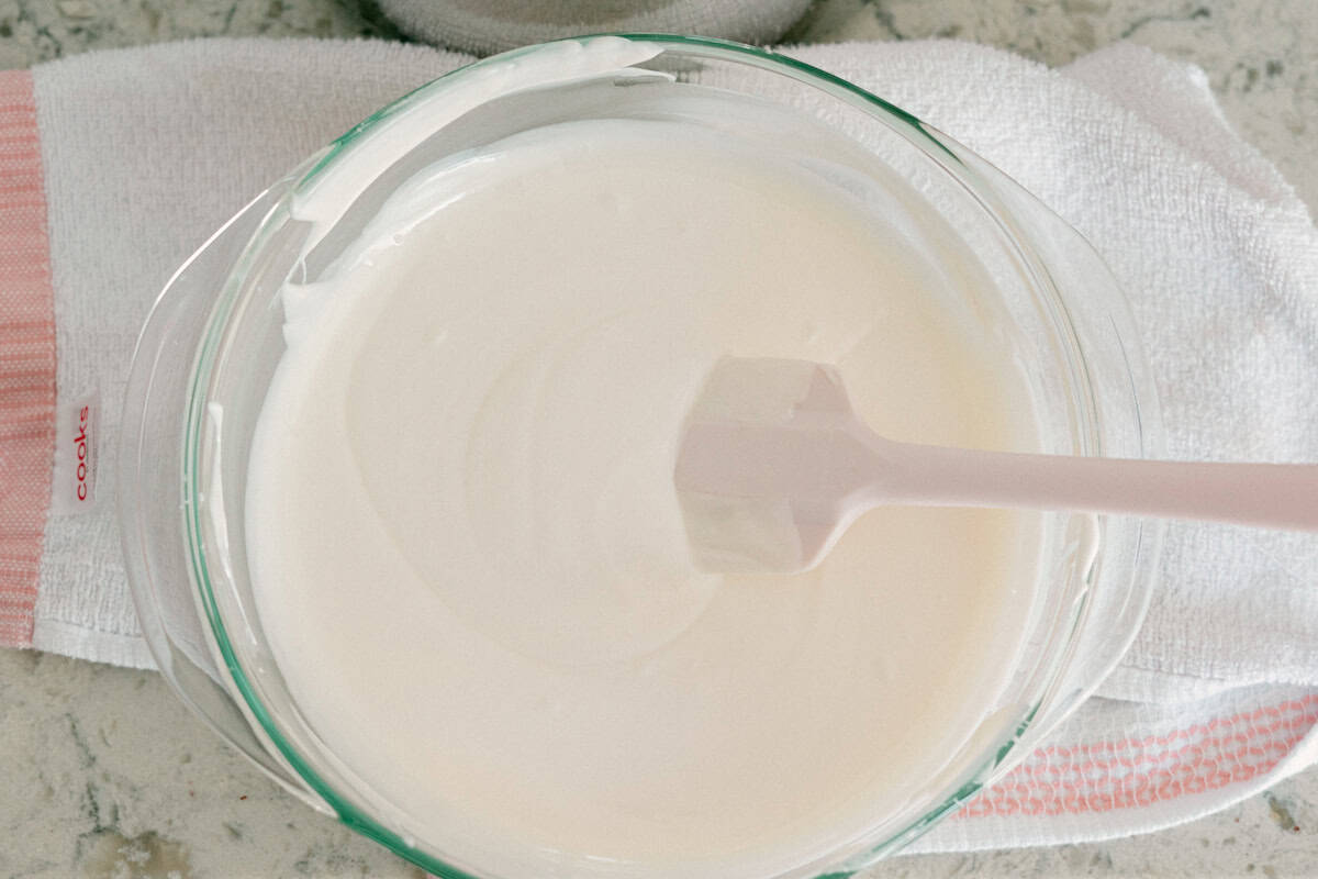 A glass bowl filled with smooth white icing or frosting, with a spatula resting inside, placed on a white towel on a countertop.