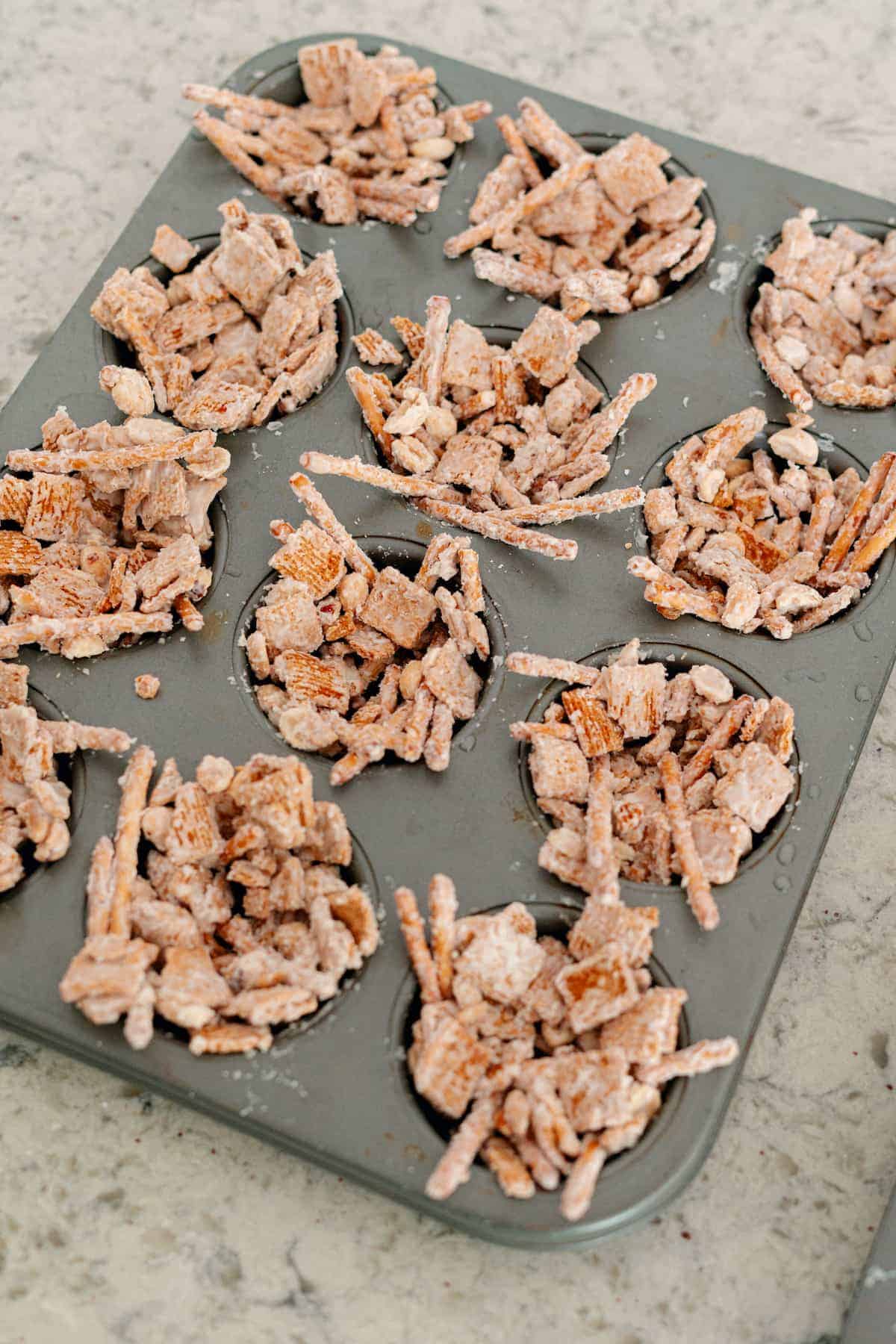 A muffin tin filled with clusters of cereal snack mix, coated in a powdered topping, placed on a kitchen countertop.