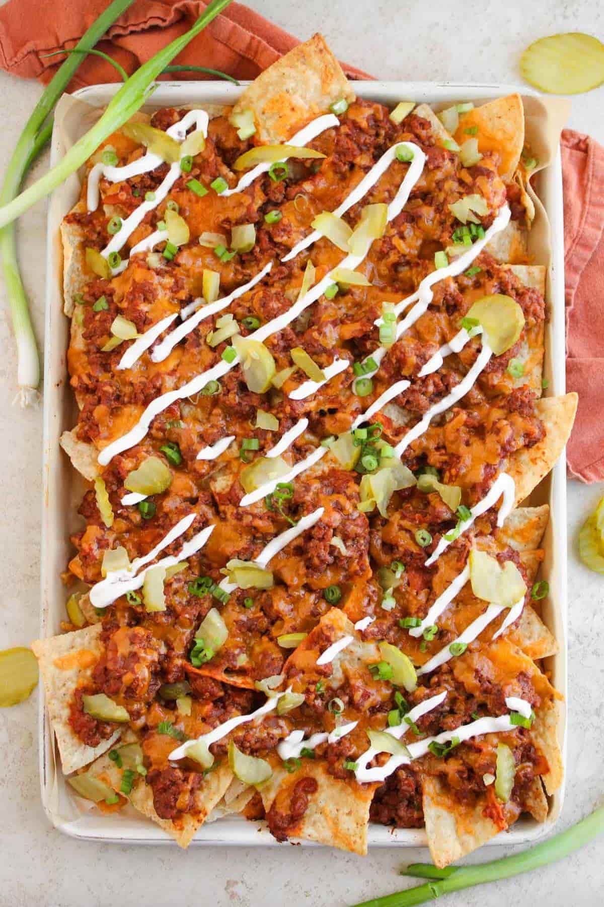 A tray of nachos topped with ground meat, melted cheese, pickles, chopped green onions, and drizzled with sour cream. Some green onions and a napkin are beside the tray.