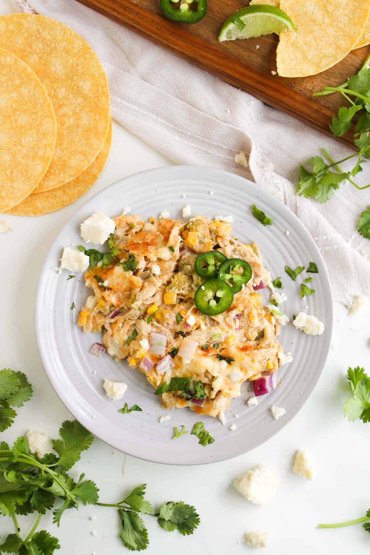 A plate of creamy chicken and corn mixture garnished with sliced jalapeños, onion, and cilantro, with corn tortillas, lime, and cheese crumbles nearby.
