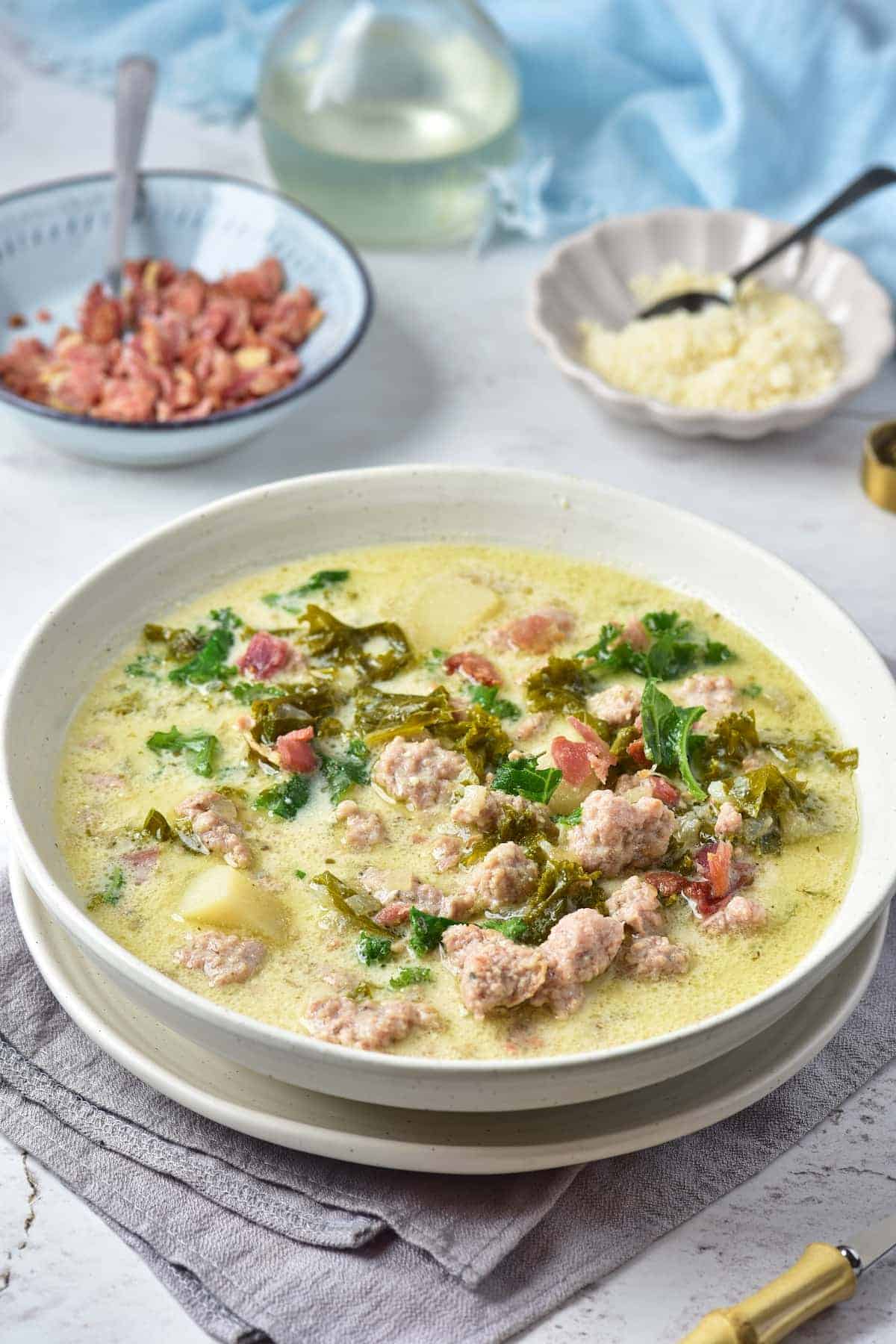 A bowl of creamy soup with meatballs, potatoes, kale, and herbs, with bowls of grated cheese and cooked bacon in the background.