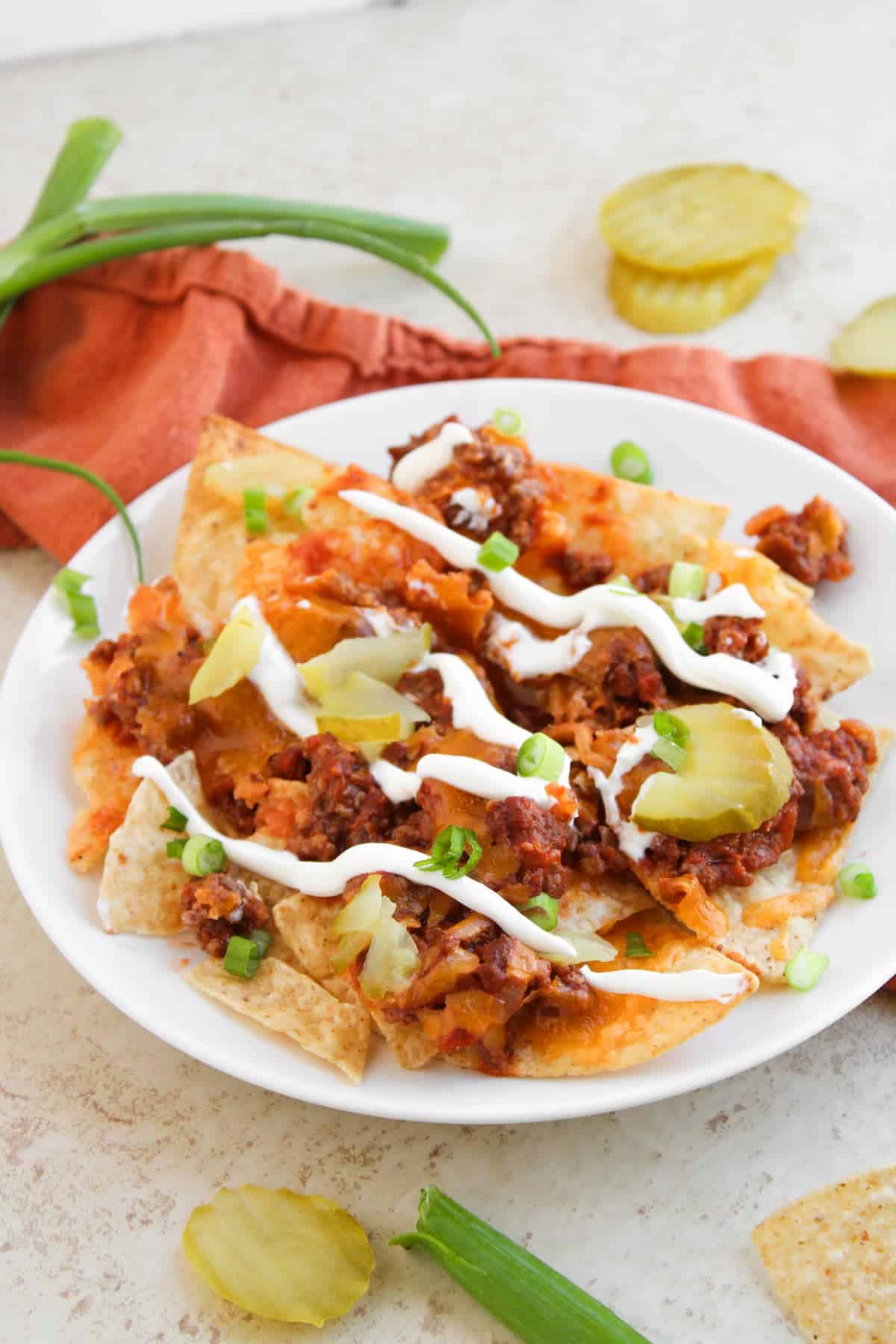 A plate of nachos topped with ground meat, melted cheese, pickle slices, sour cream, and chopped green onions sits on a light surface with scattered pickles and green onions nearby.