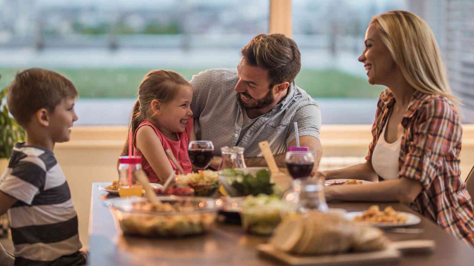 A family of four sits at a dining table, sharing a meal and smiling at each other in a brightly lit room.