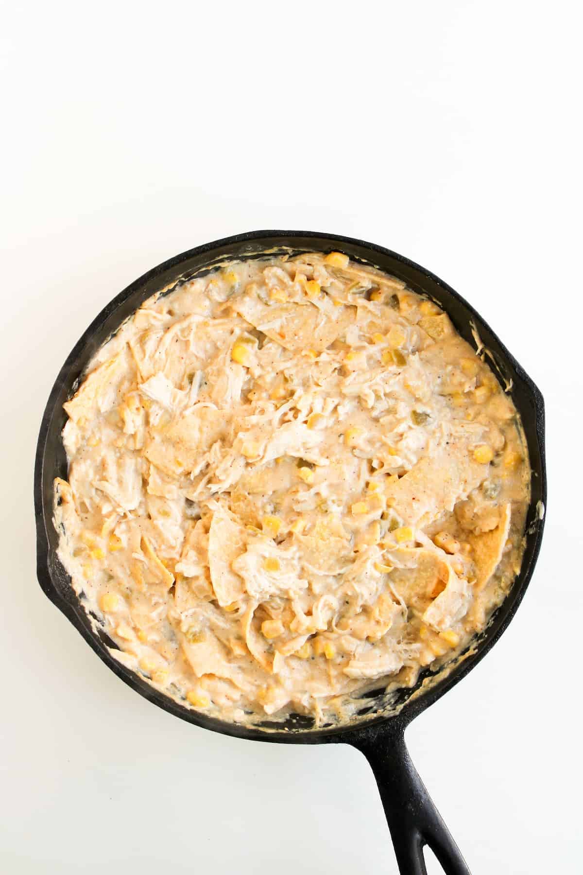 A cast iron skillet filled with a creamy mixture of corn, tortilla chips, cheese, corn, and other ingredients on a white background.