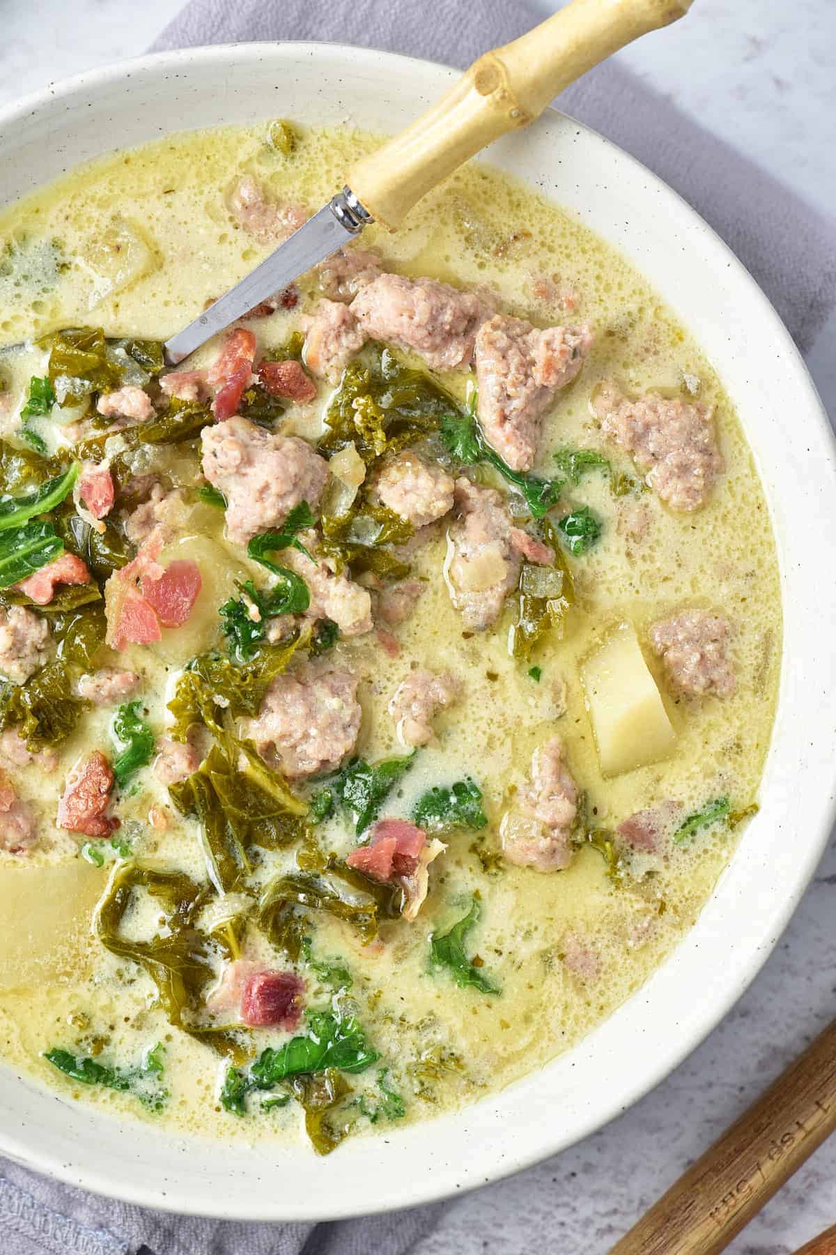 A bowl of creamy soup with sausage, potatoes, kale, tomatoes, and a wooden-handled spoon resting on the edge.