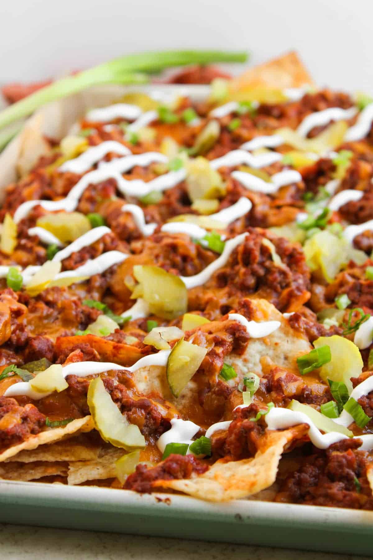 A tray of nachos topped with ground beef, melted cheese, sliced pickles, chopped green onions, and drizzled with sour cream.