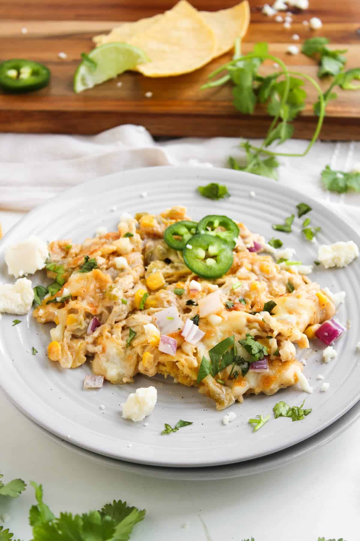 A plate of cheesy Mexican casserole topped with sliced jalapeños, red onion, cilantro, and crumbled cheese, with tortilla chips and lime wedges in the background.