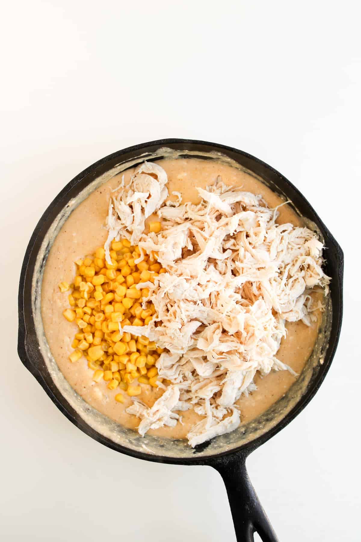 A cast iron skillet filled with creamy mixture, topped with shredded chicken and corn, sits on a white background.