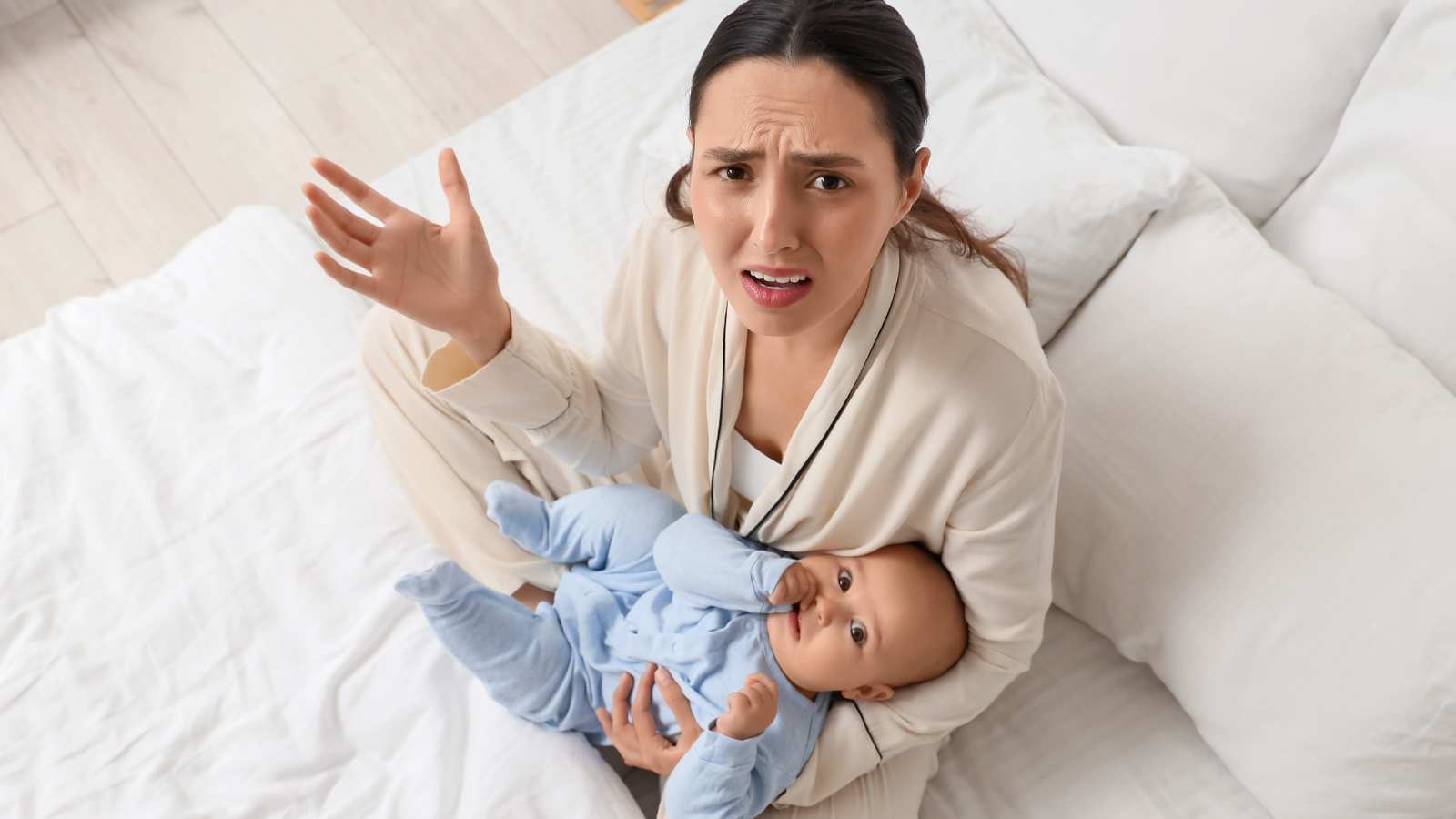 A woman sits on a bed holding a baby in her lap, looking up with a confused or frustrated expression and one hand raised.