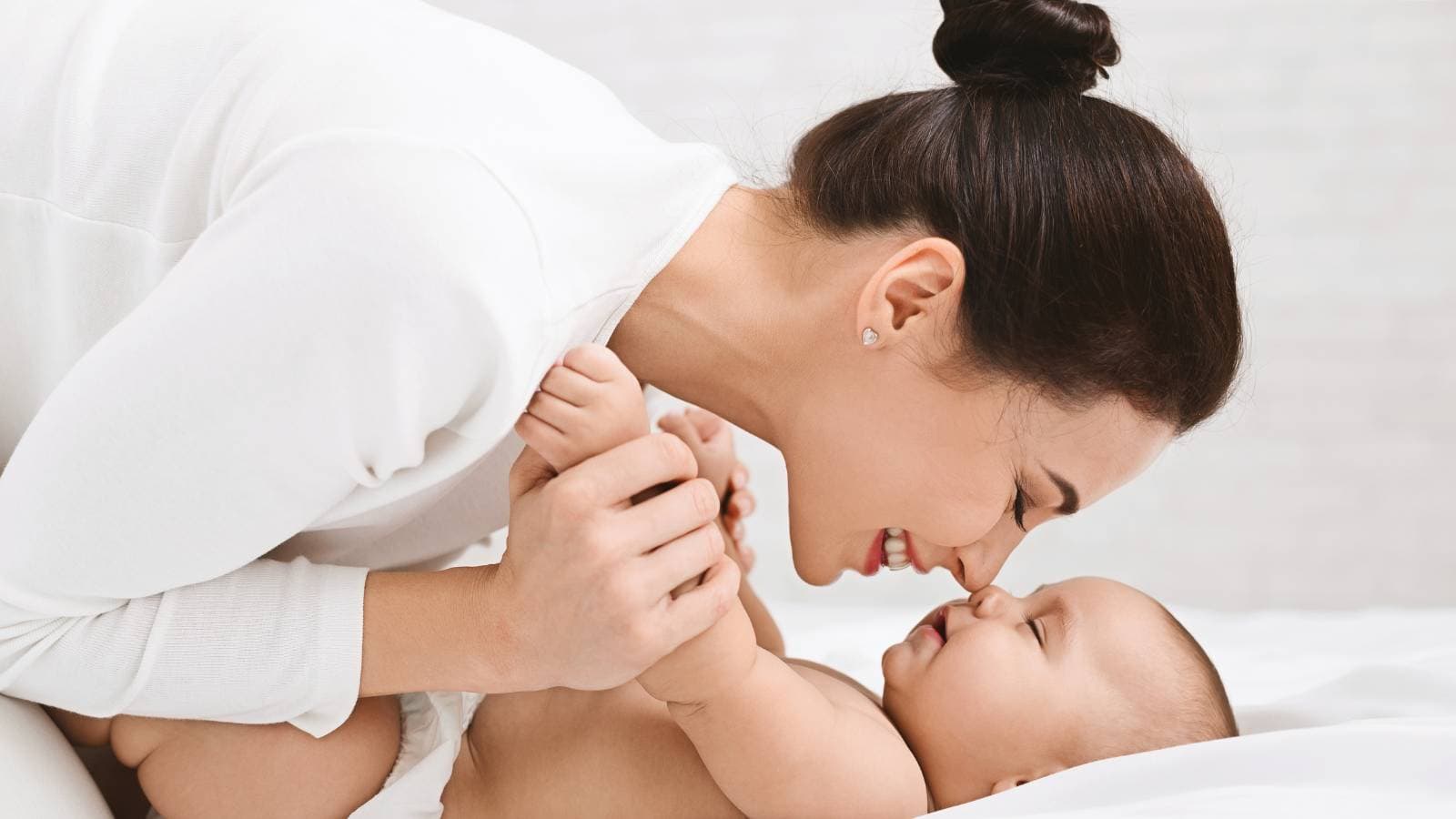A woman leans down and touches noses with a smiling baby while holding the baby's hands on a white bed.