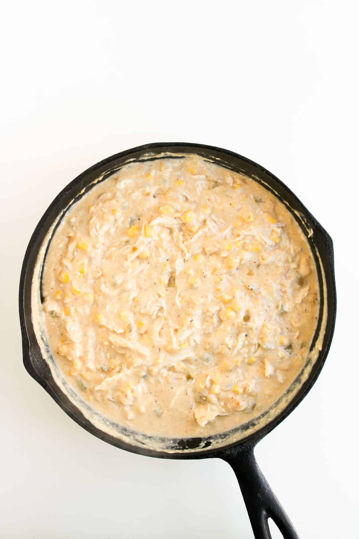 A cast iron skillet filled with a creamy mixture containing shredded chicken, corn, and sauce, viewed from above on a white background.