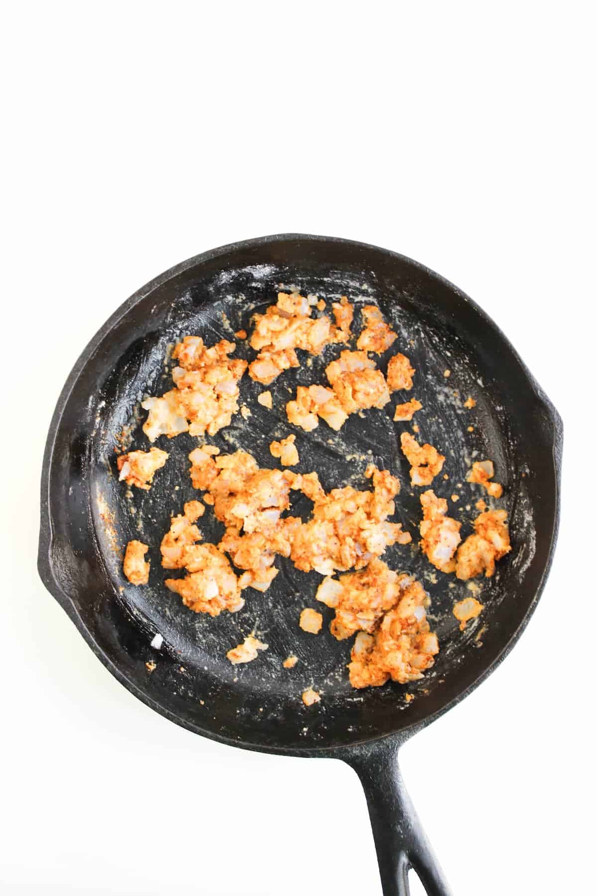 A black cast iron skillet containing partially cooked ground meat with seasonings and small bits of onion, on a white background.