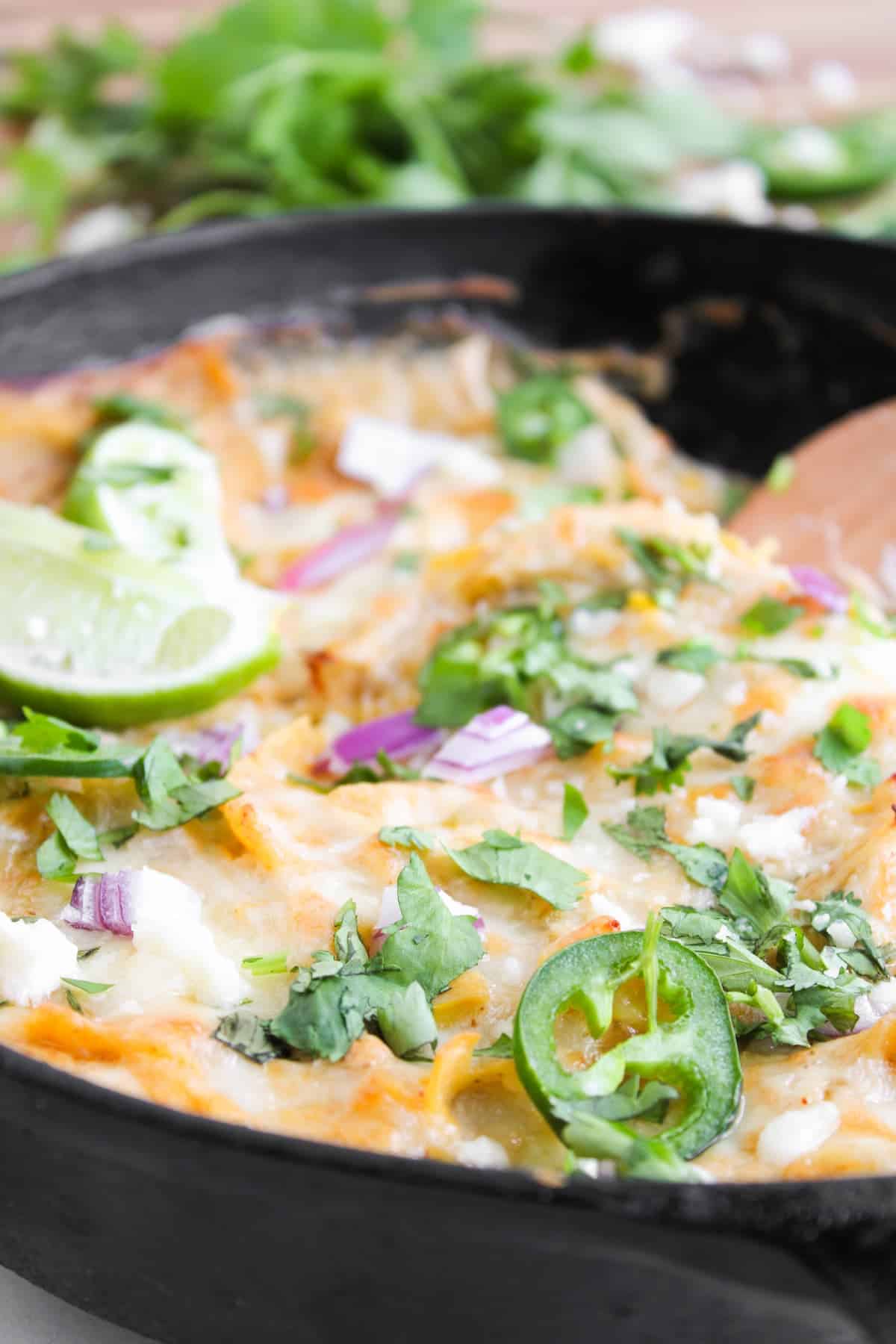 A close-up of a baked cheesy white chicken enchilada in a skillet, topped with sliced jalapeño, cilantro, red onion, lime, and crumbled cheese.