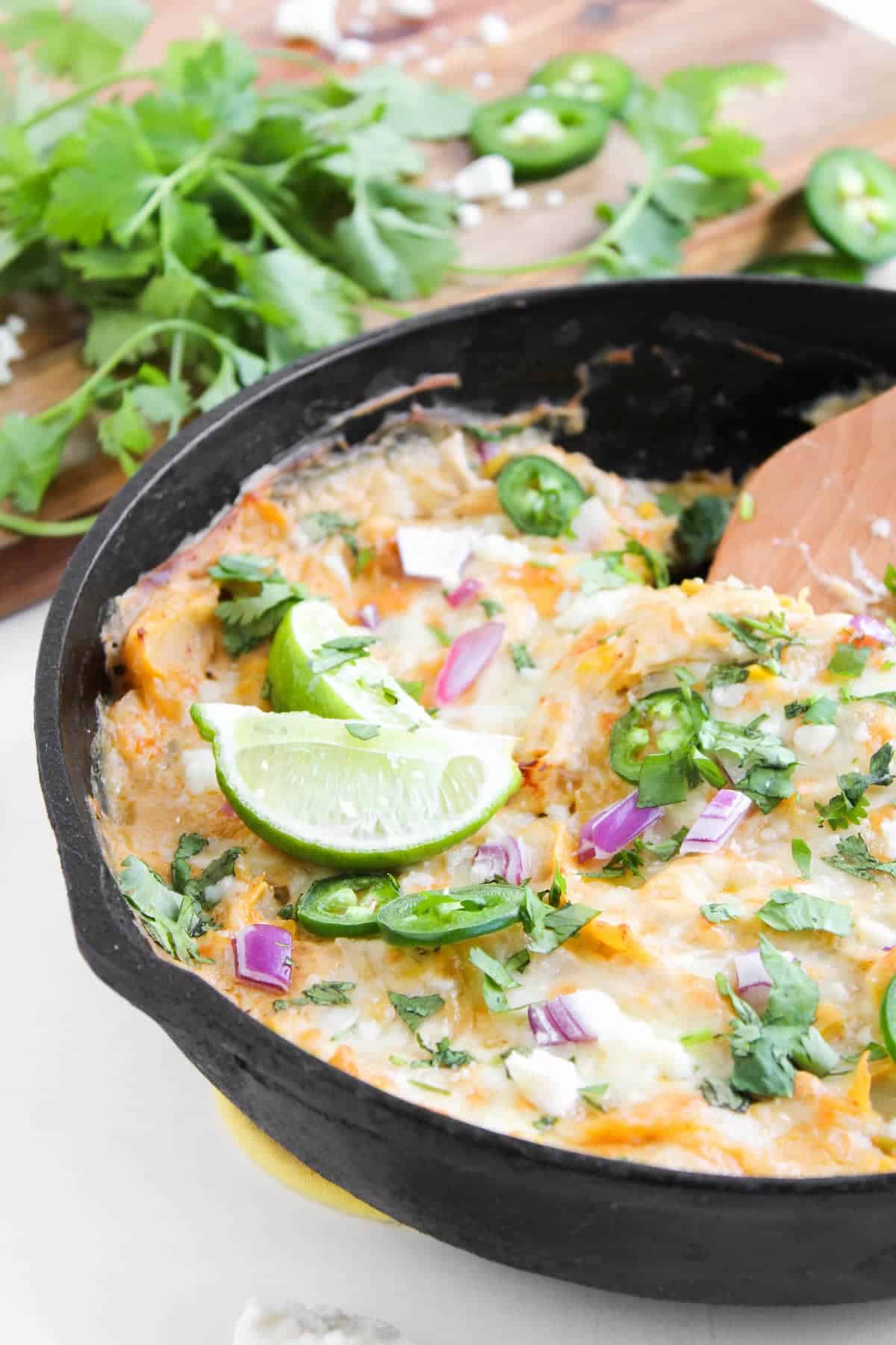 A cast iron skillet filled with cheesy baked dip, topped with sliced jalapeños, red onion, cilantro, and lime wedges. Fresh cilantro and jalapeño slices are on a cutting board in the background.