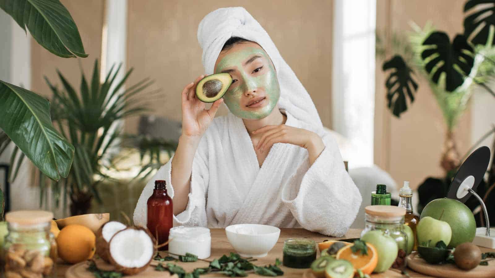 Woman in a white bathrobe with a green face mask holds half an avocado over one eye, seated at a table with fruits and skincare products.