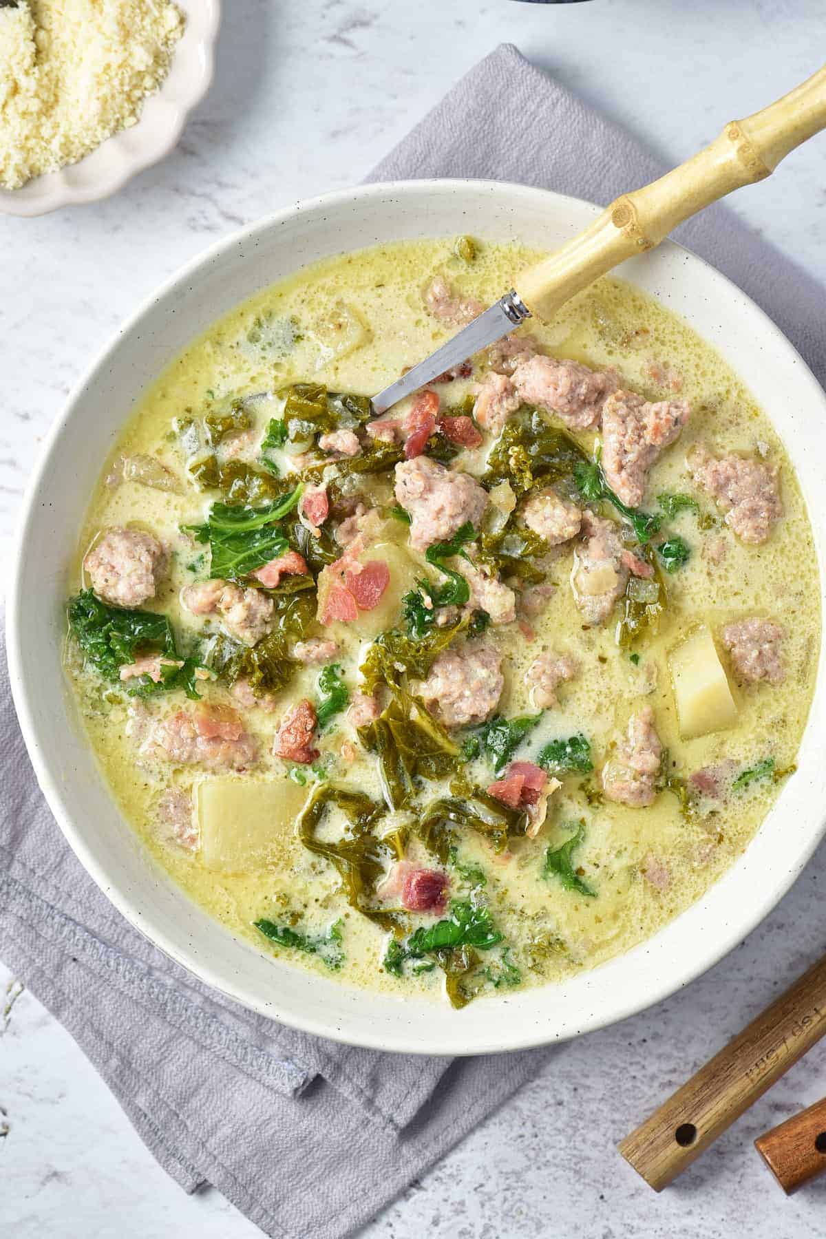 A bowl of creamy soup with sausage, potatoes, kale, and tomatoes, served with a spoon on a gray napkin.