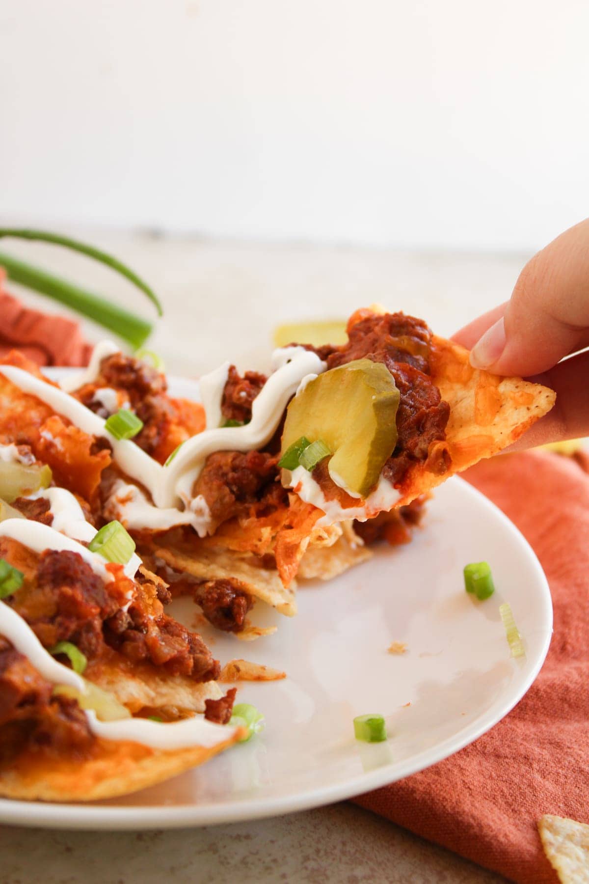 A hand lifts a nacho chip topped with ground beef, pickles, green onions, and sour cream from a plate of loaded nachos.