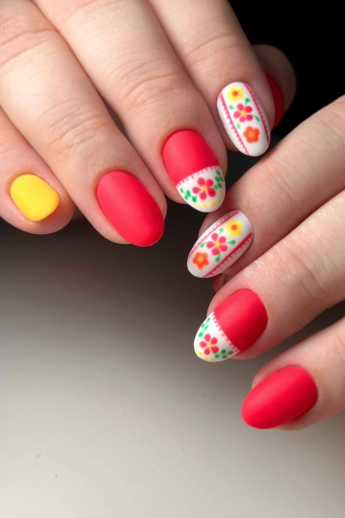 A hand with matte red and yellow nails, and two accent nails featuring colorful floral patterns on a white background.