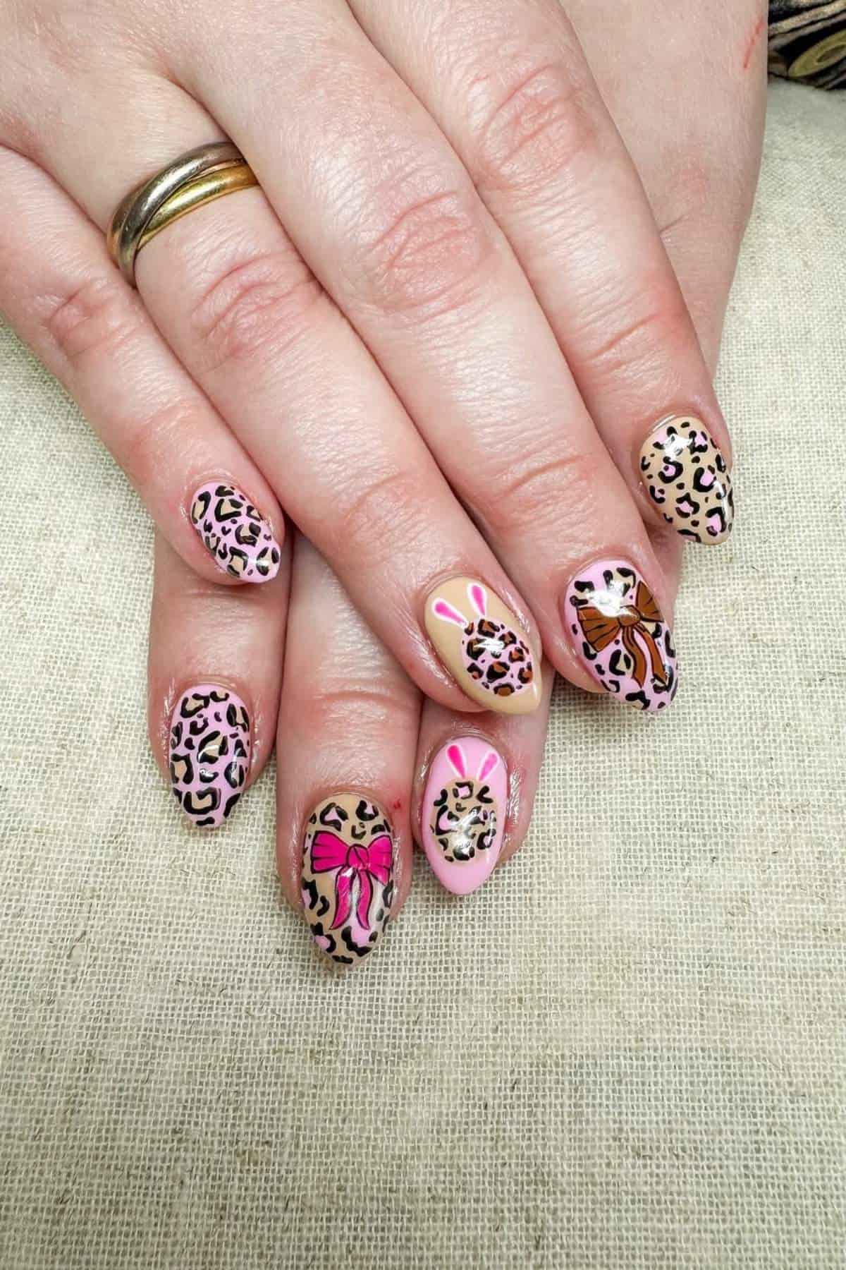 Hands with almond-shaped nails featuring pink leopard print, pink bows, and stylized bunny designs on a light fabric surface.